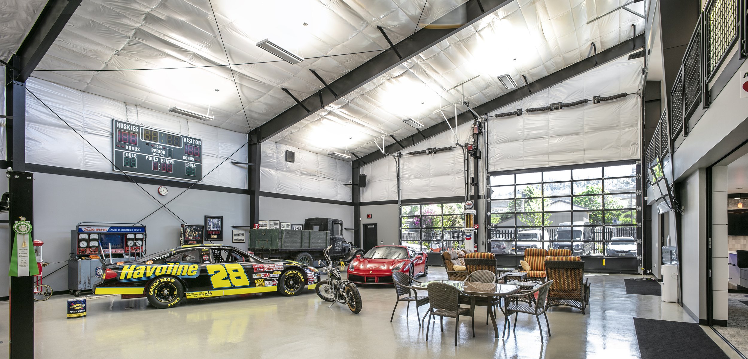 Inside a modern automotive garage with a race car, a classic sports car, a bicycle, and seating area near large windows.