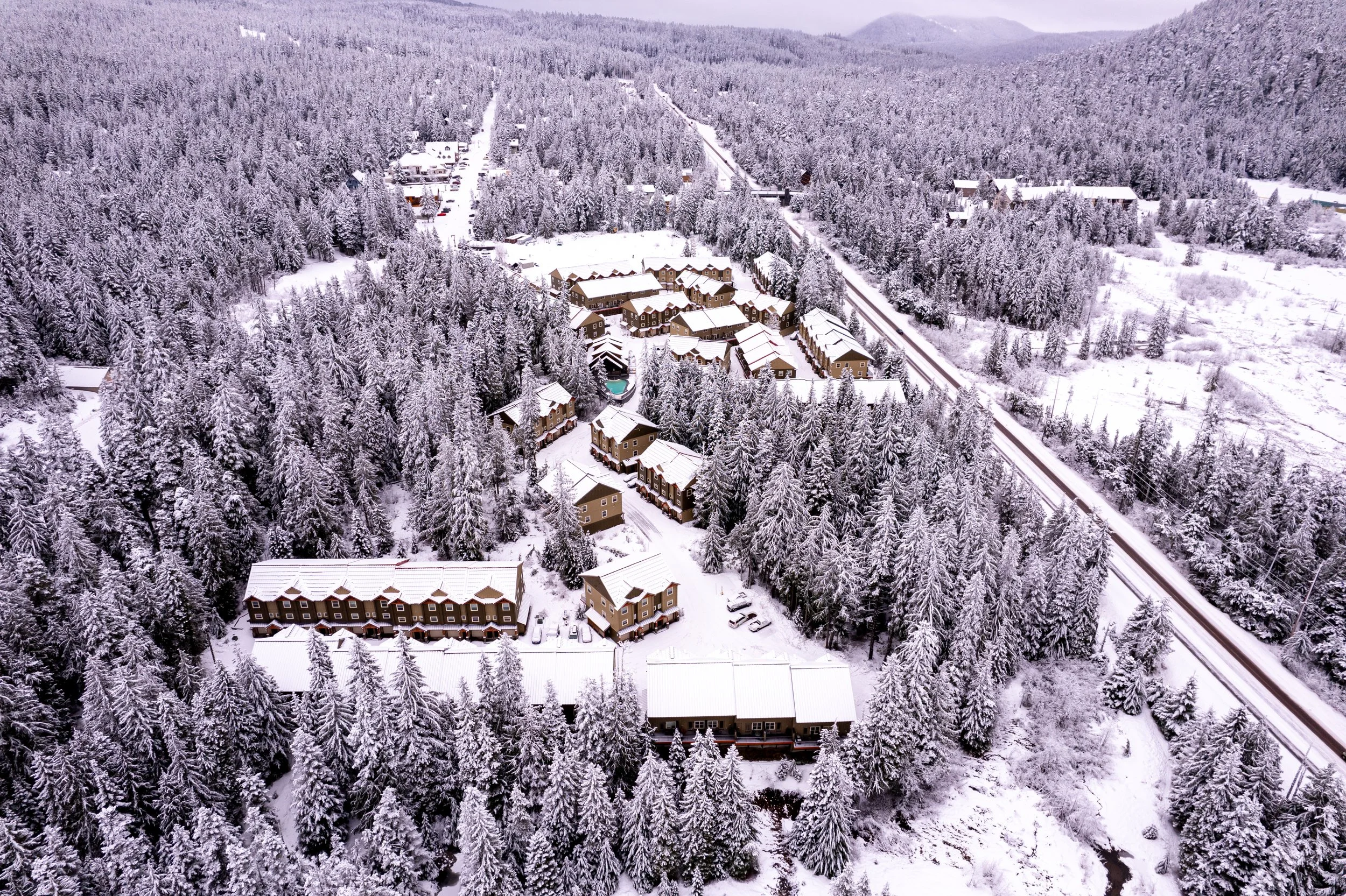 An aerial view of a snow-covered mountain village surrounded by dense forest, with buildings, roads, and train tracks visible.