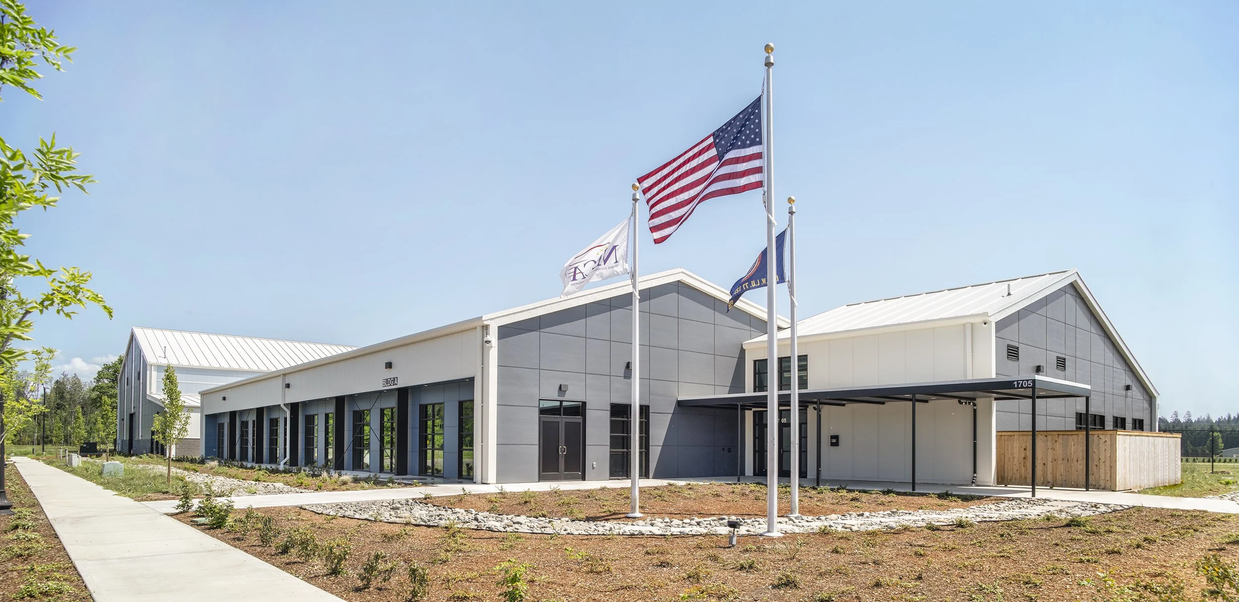 Modern gray building with three flagpoles in front, displaying the American flag, a white flag, and a blue flag, under a clear blue sky. Sidewalk and landscaped area with young plants and small trees surround the building.