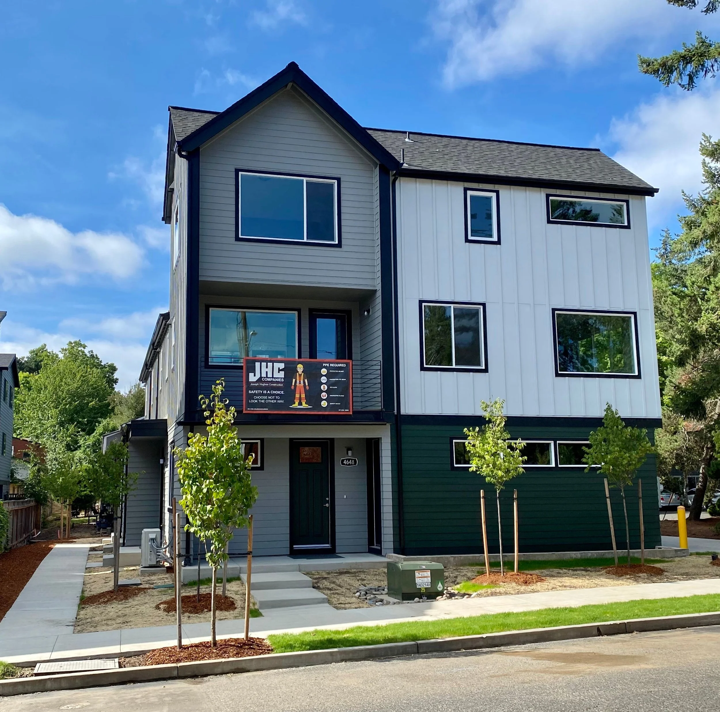 New multi-story residential building with gray and white exterior, black window frames, and small front yard with young trees and sidewalk.