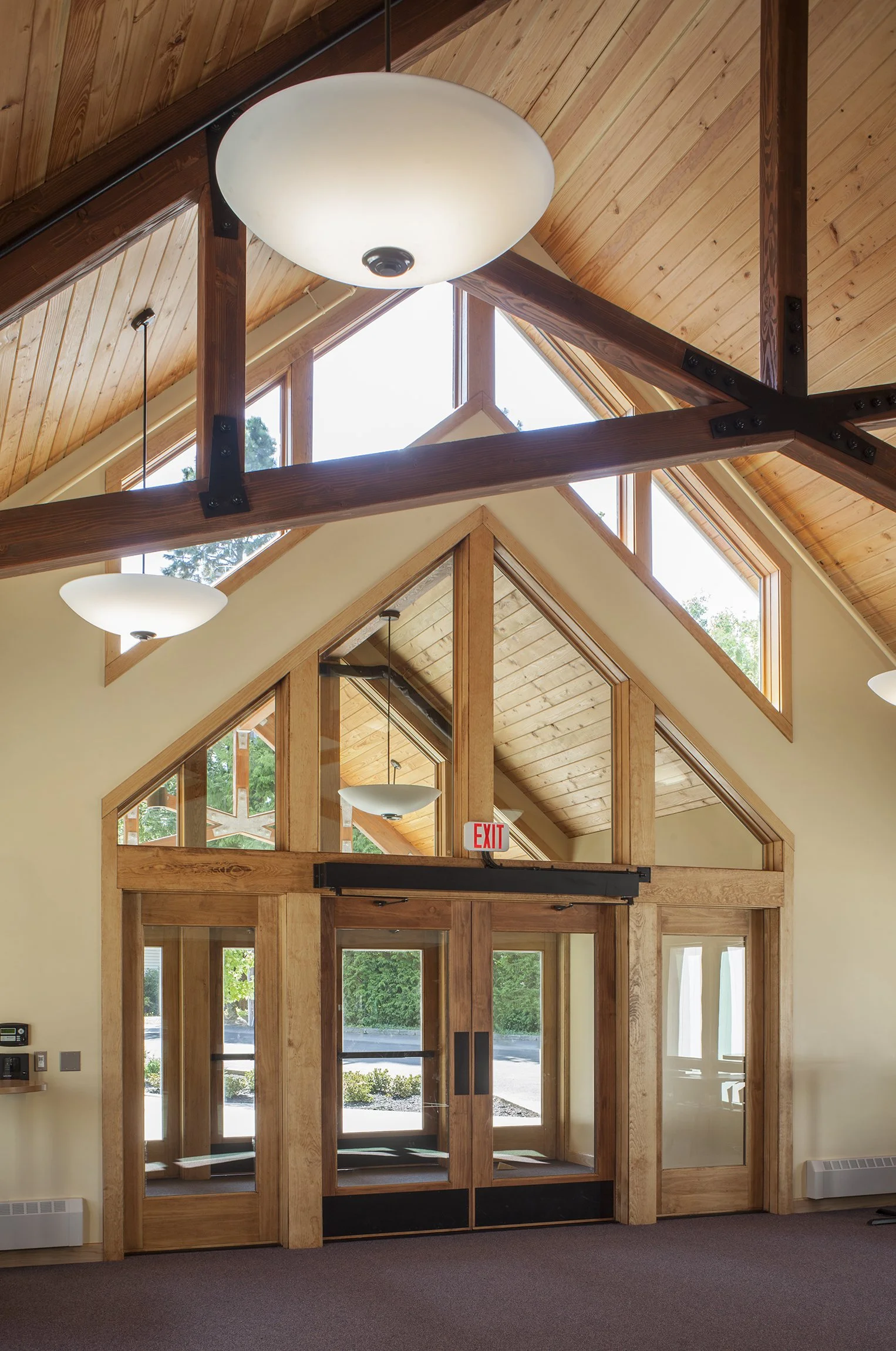 Interior view of a building with a high vaulted ceiling, wooden beams, large triangular windows, hanging light fixtures, glass doors, and an exit sign.