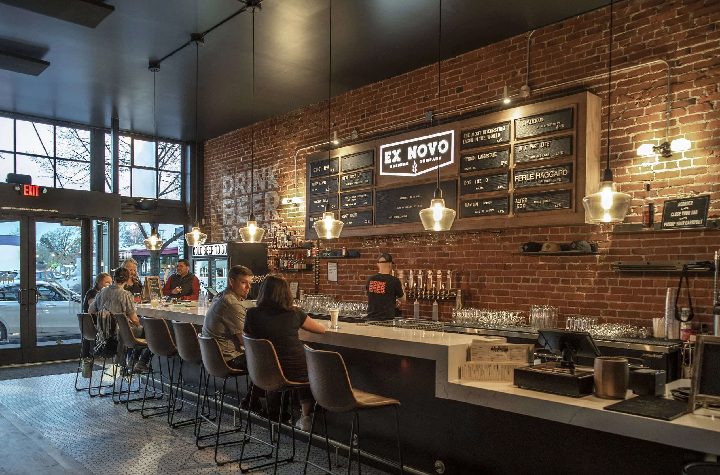 Inside a modern bar with a brick wall, a white marble counter, hanging pendant lights, a chalkboard menu, and people sitting at the bar drinking and talking.