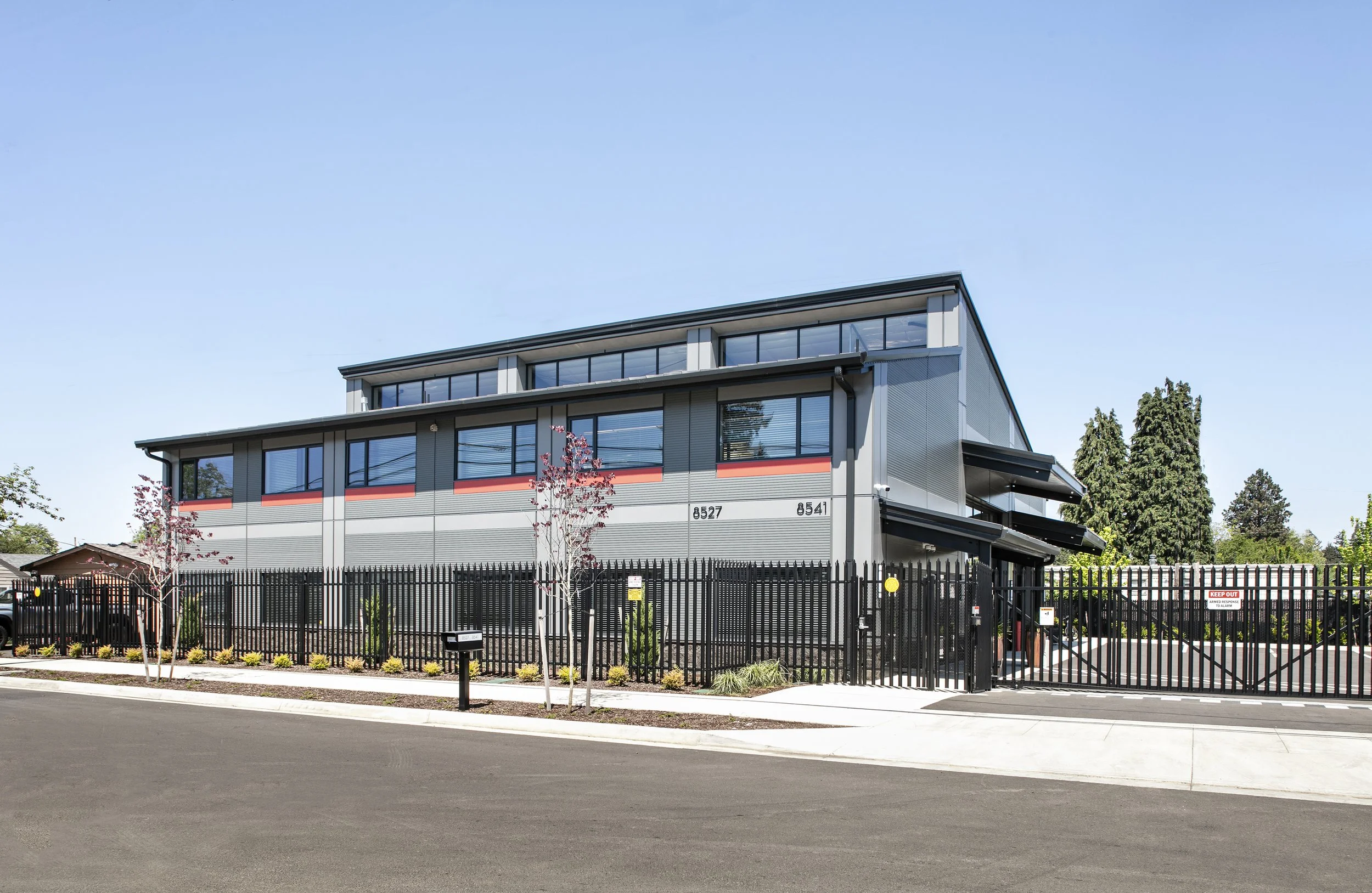 A modern, multi-story building with large glass windows, a black metal fence, and a small parking area with some cars and trees along the street.