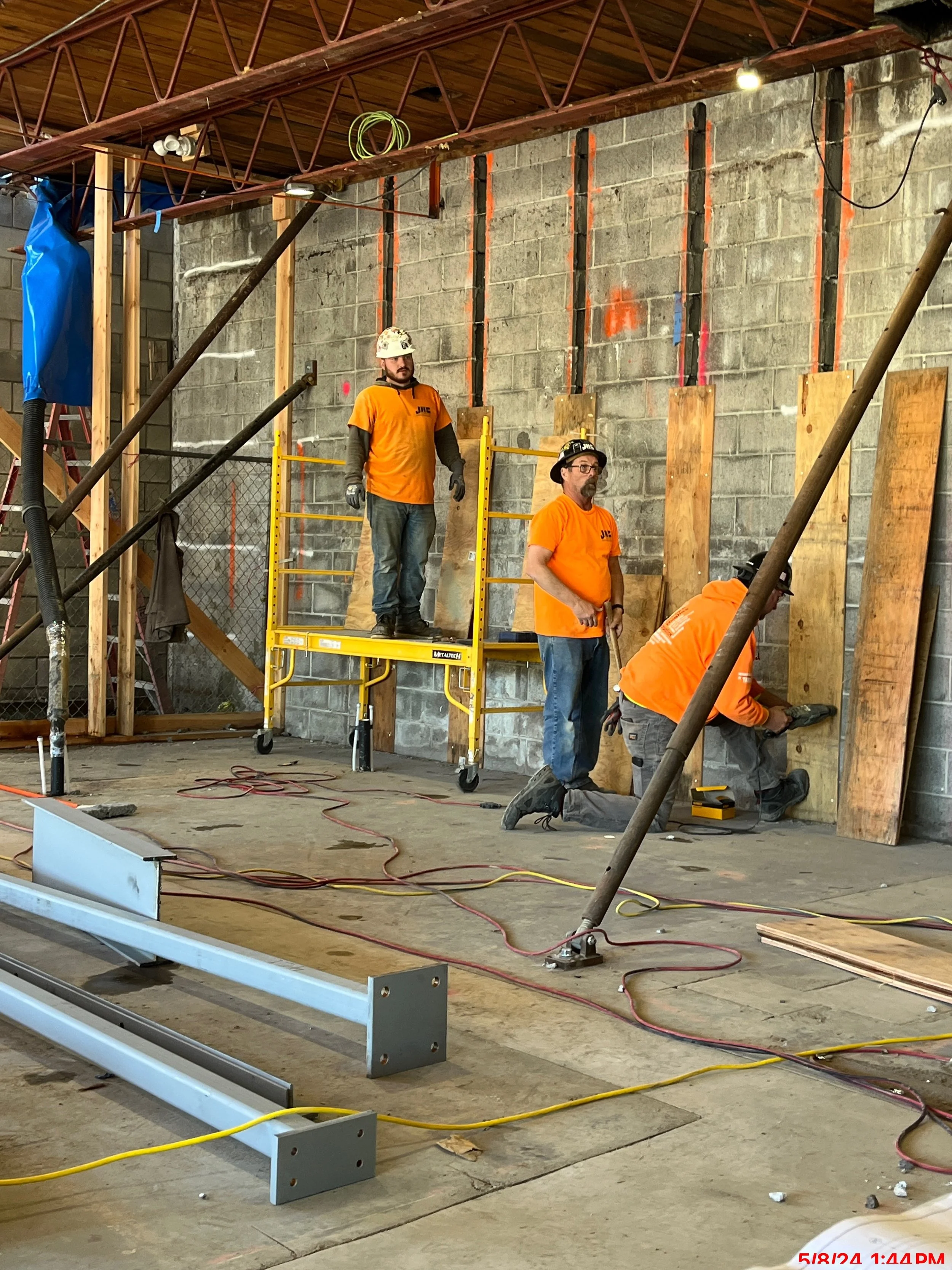 Construction workers in orange shirts and helmets working on a wall inside a building, with scaffolding and construction tools around.