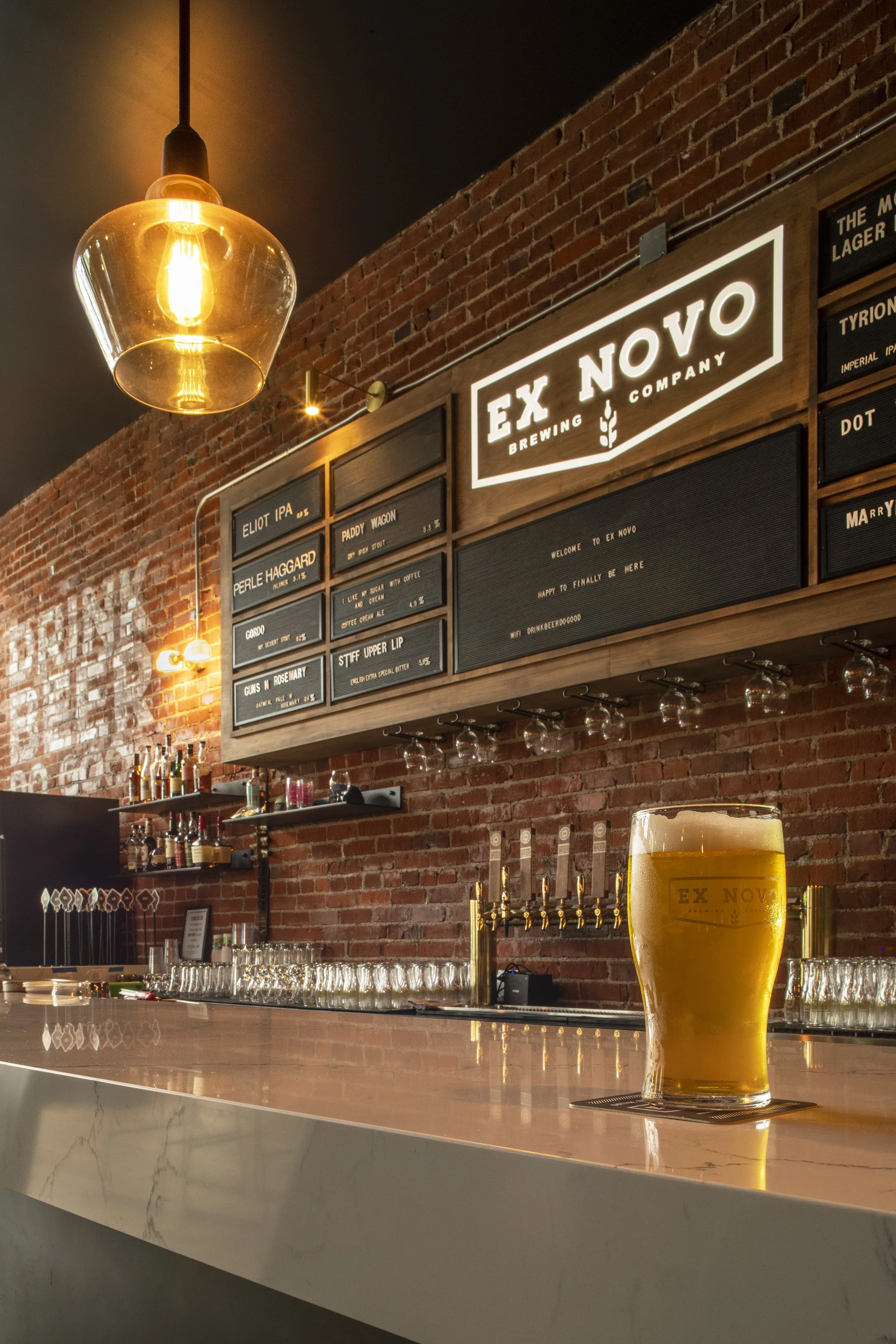 A bar with a glass of beer on the counter, exposed brick wall, hanging light fixture, and illuminated sign reading 'EX NOVO Brewing Company'.