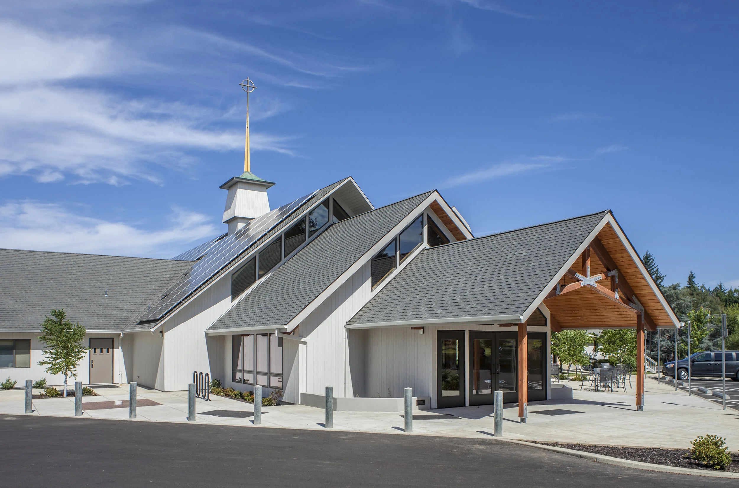 Modern church building with solar panels on roof, wooden entrance, and outdoor seating area under a covered porch, against a blue sky with clouds.