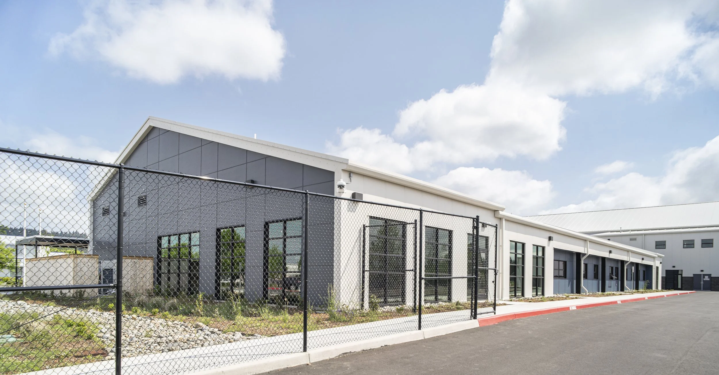 A modern commercial building with gray and white exterior, large windows, surrounded by a black chain-link fence and parking lot under a partly cloudy sky.