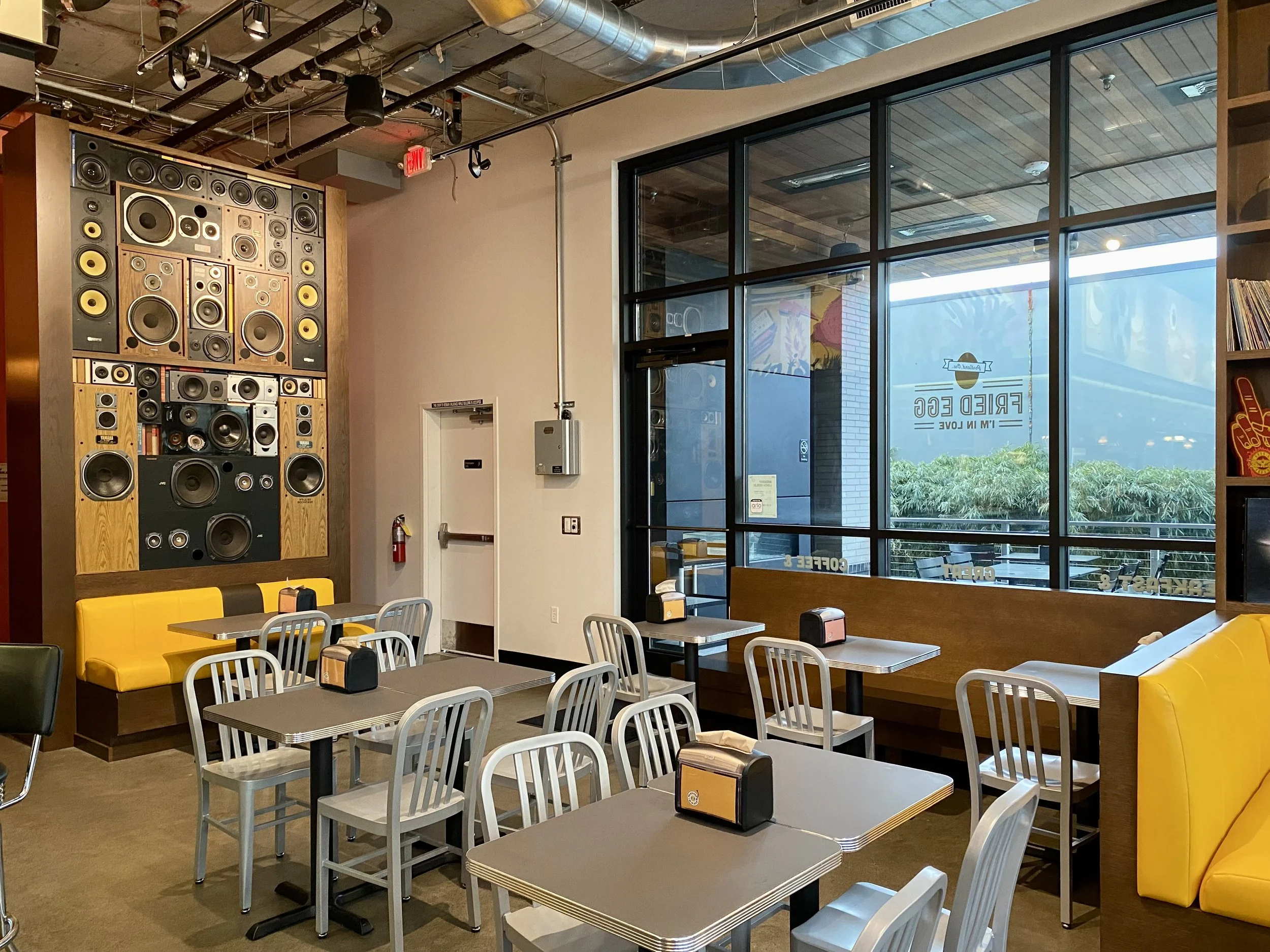 An interior of a restaurant or cafe with multiple tables and chairs, a large black and yellow speaker wall, large windows with a view outside, and a yellow cushioned seating area.