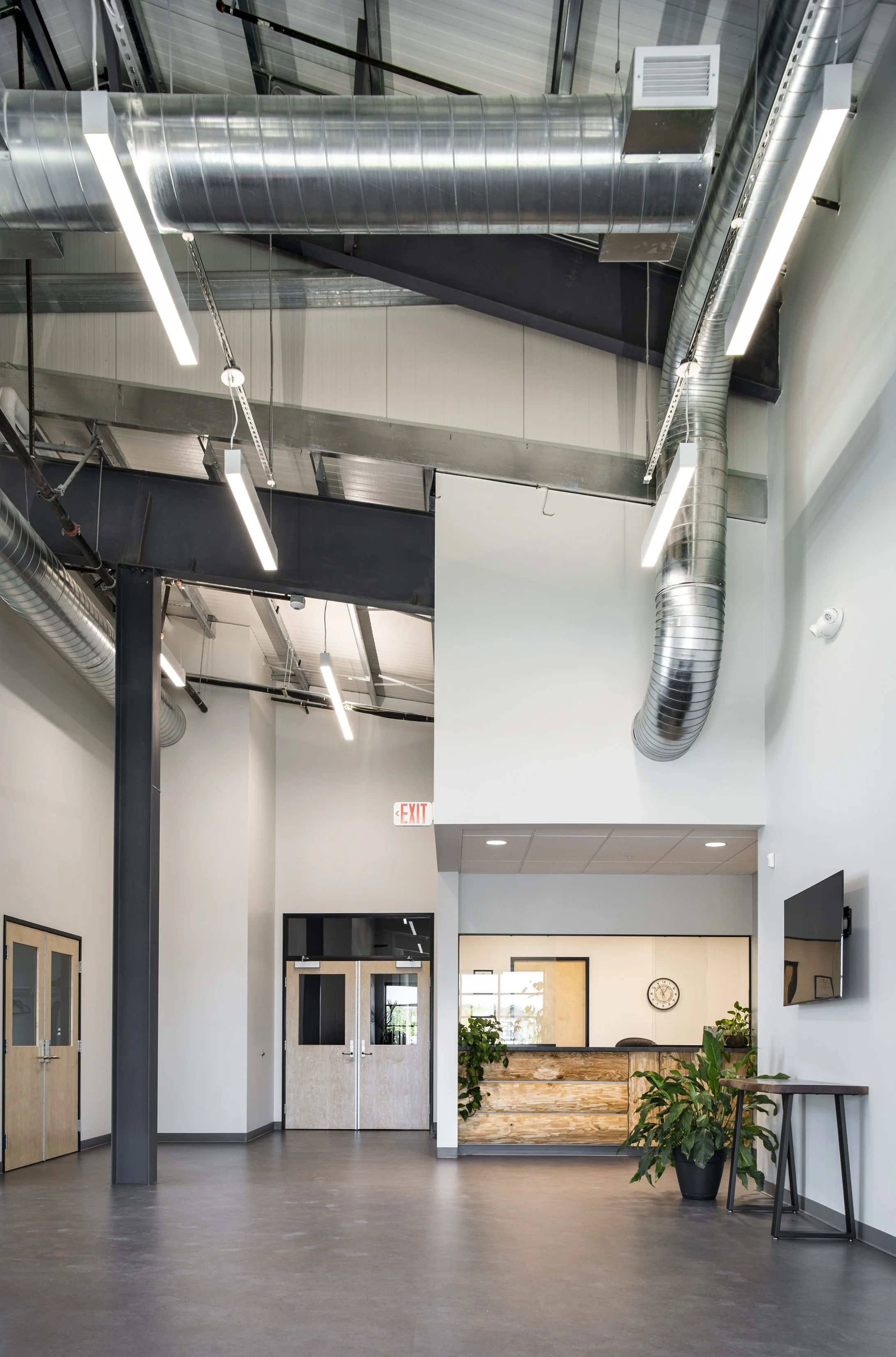 An empty modern lobby with a reception desk, potted plants, a clock, a mounted TV, and an exit sign, featuring exposed ductwork and industrial-style lighting.