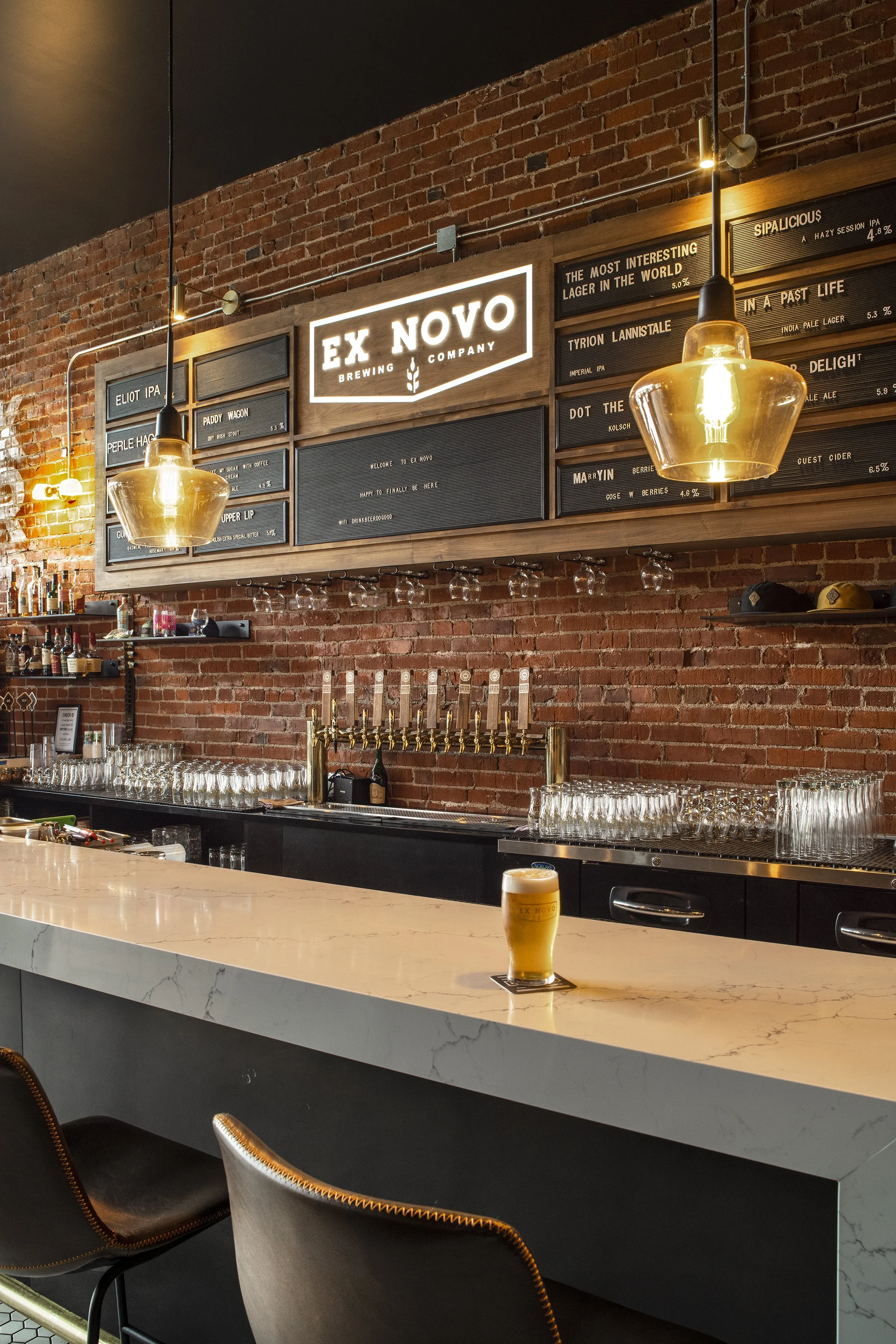 A bar with a white marble countertop and black cabinets. A glass of beer with a coaster sits on the counter. Behind the bar is a brick wall with a liquor shelf, beer taps, glassware, and signs displaying beer names and descriptions. Two pendant light