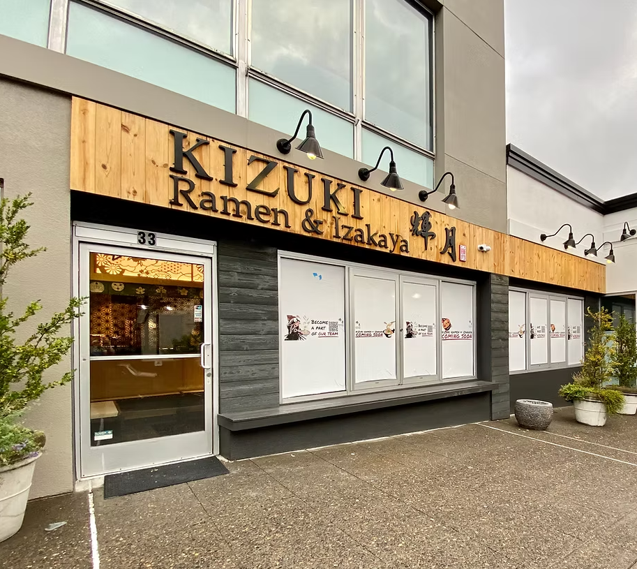 Exterior view of Kizuki Ramen & Izakaya restaurant, showing glass windows with posters, a wooden sign with the restaurant's name, and potted plants outside on a rainy day.