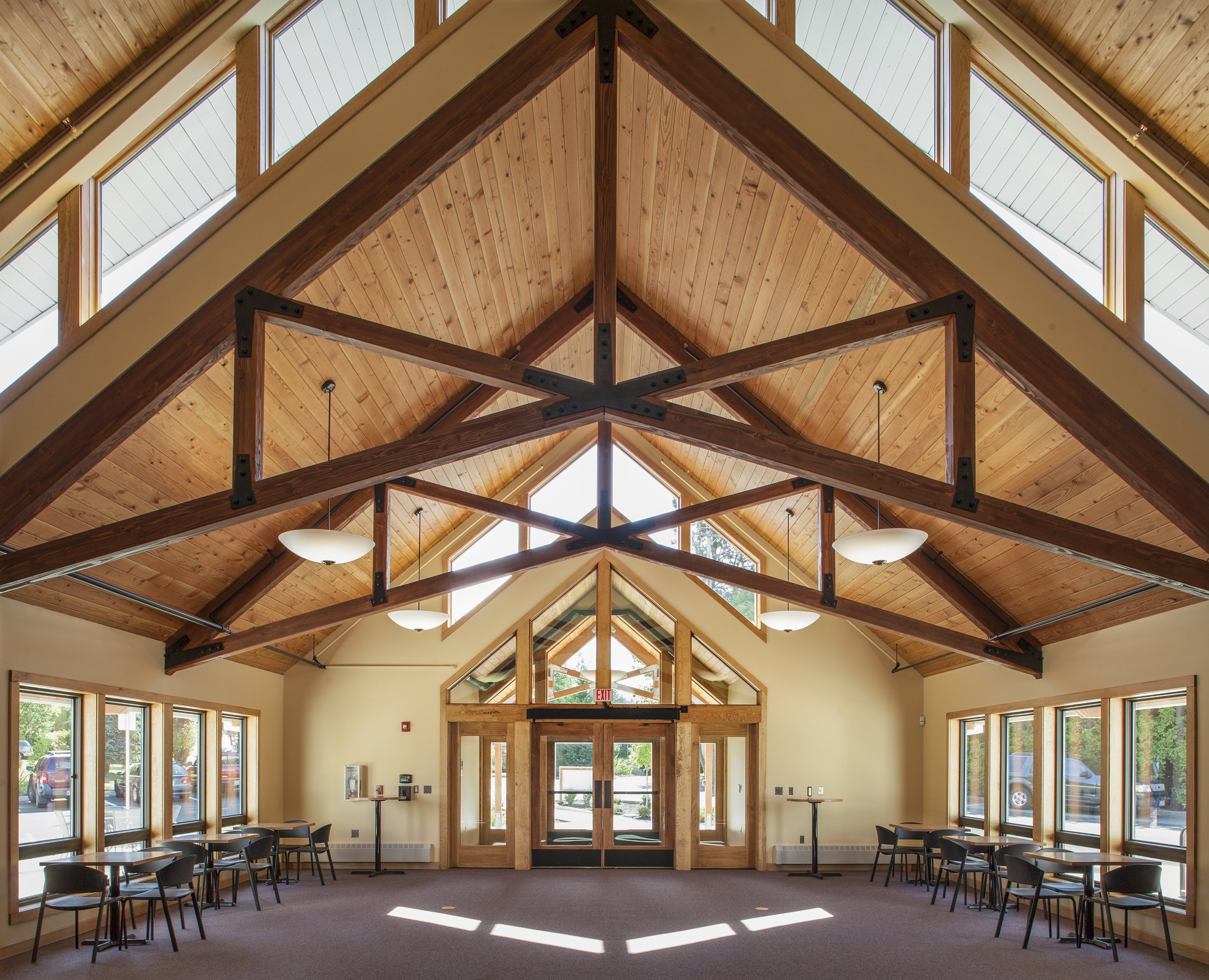 Interior of a spacious room with a high, vaulted wooden ceiling, large windows on all sides, and chairs arranged along the walls. There are wooden beams and modern hanging light fixtures.