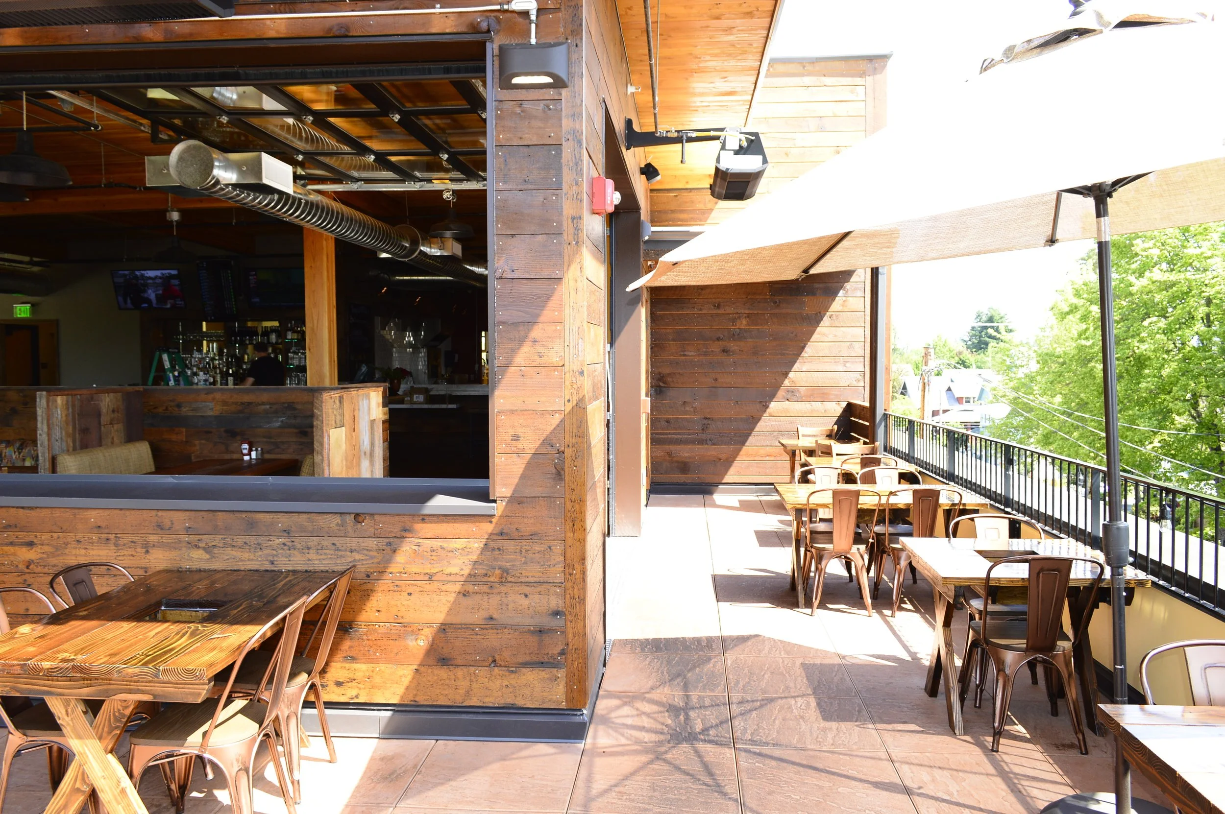 Partially enclosed outdoor patio with wooden tables and chairs, large patio umbrella, and view of green trees and neighboring buildings.