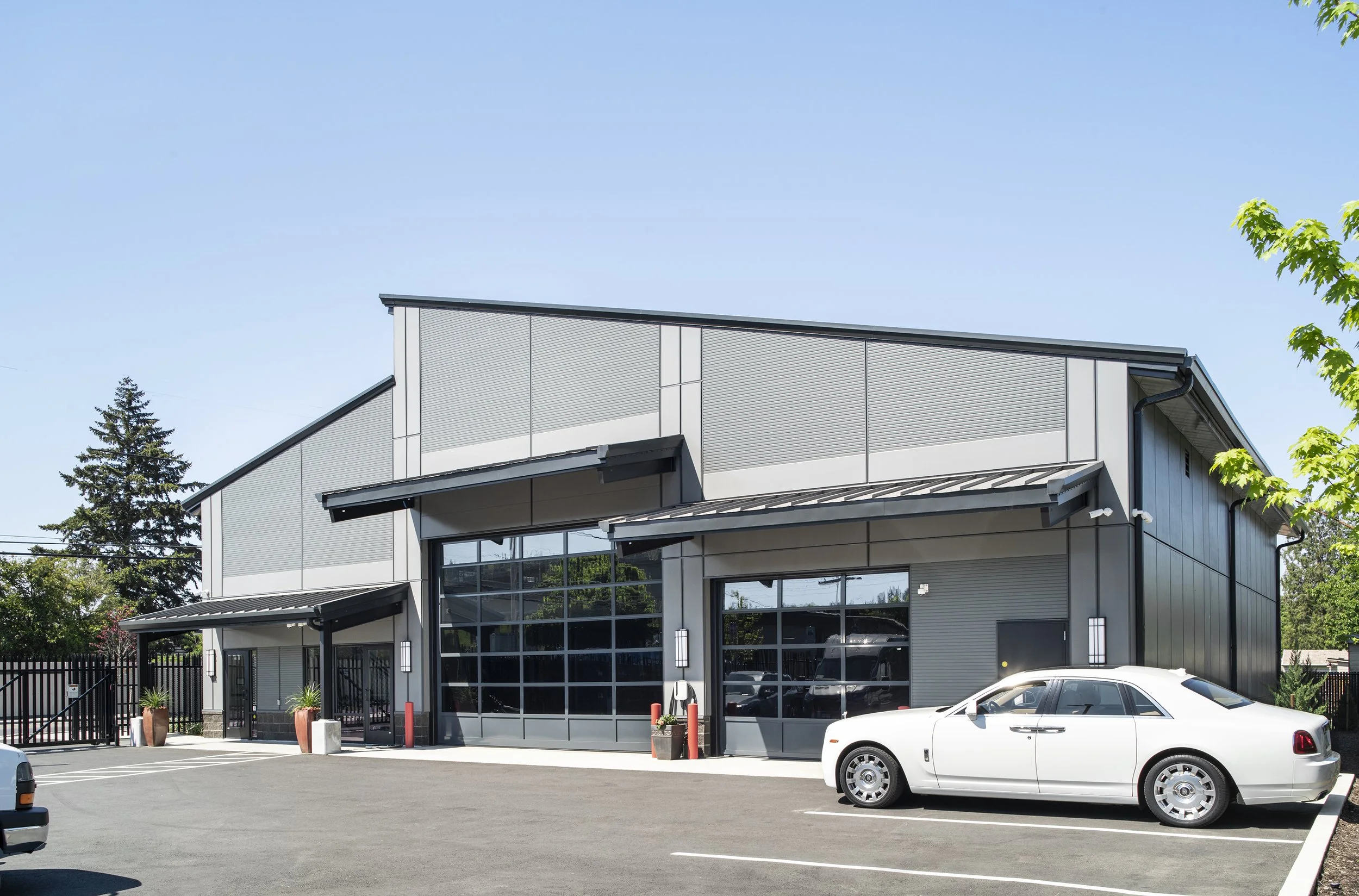 Modern commercial building with large glass garage doors and a white luxury car parked in front, surrounded by parking spaces and some green trees.