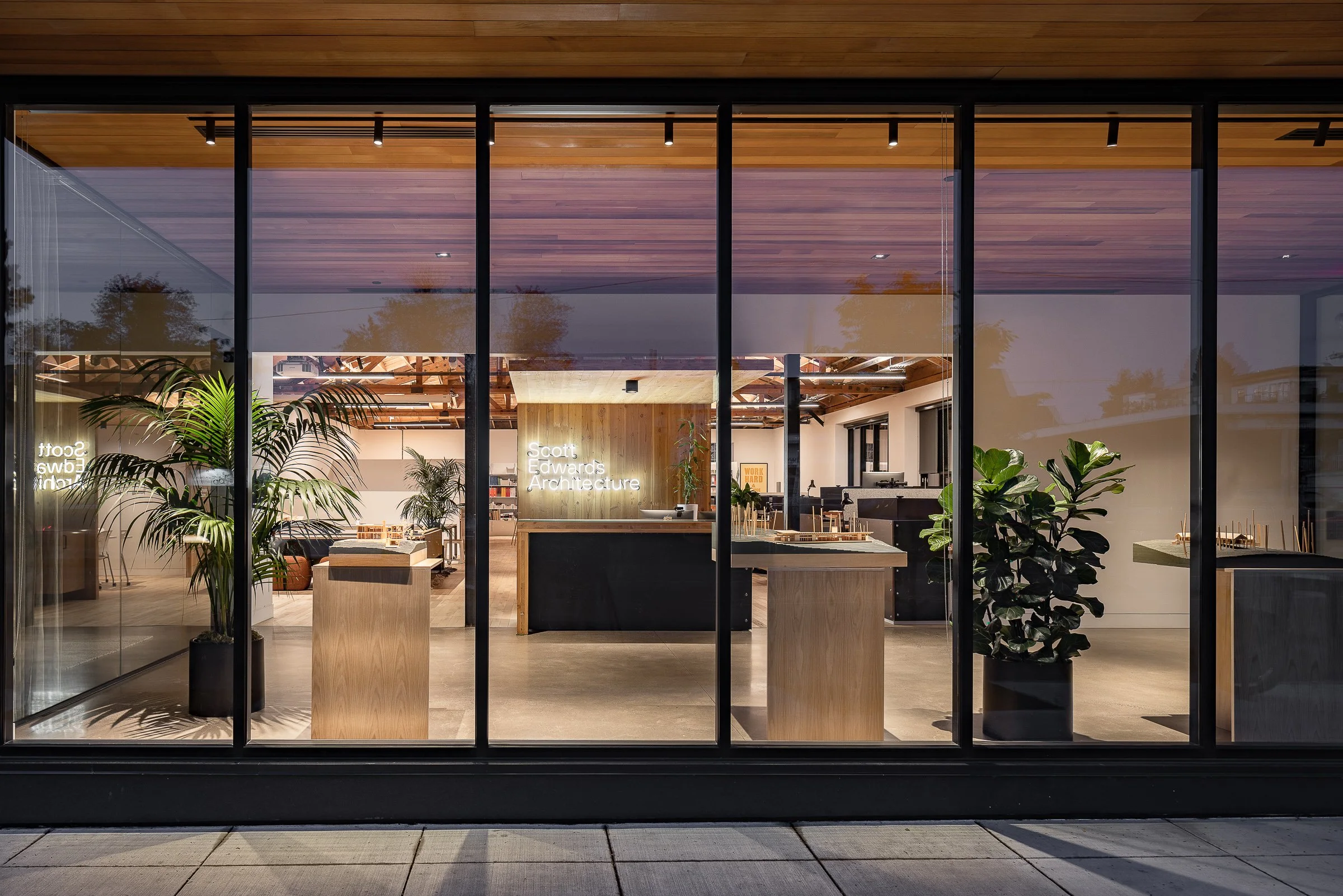 Interior of an architecture office with plants and model displays, visible through large glass windows at dusk.