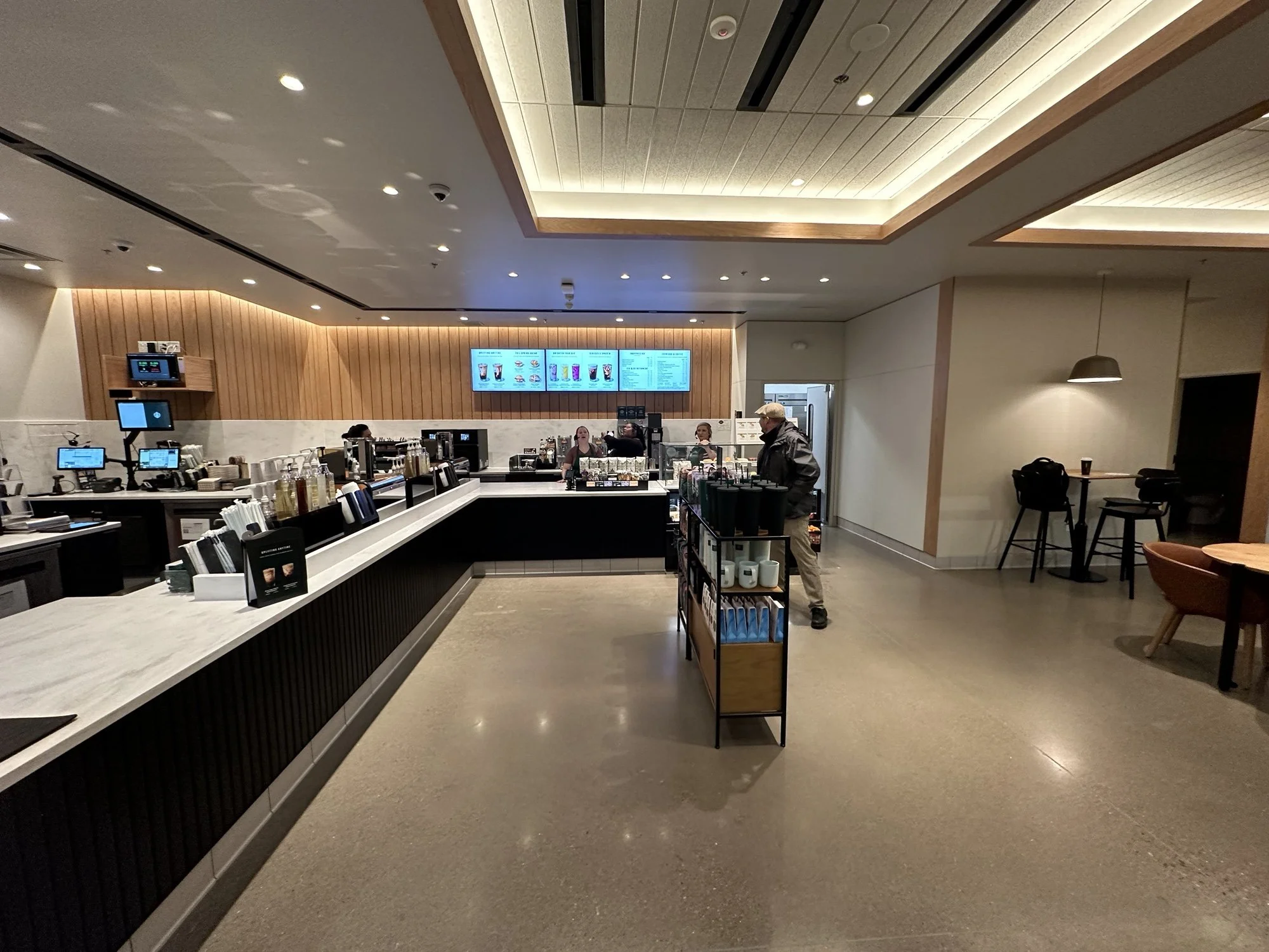 Interior of a modern cafe or coffee shop with a counter and digital menus on the wall, a few customers, and baristas behind the counter preparing drinks.