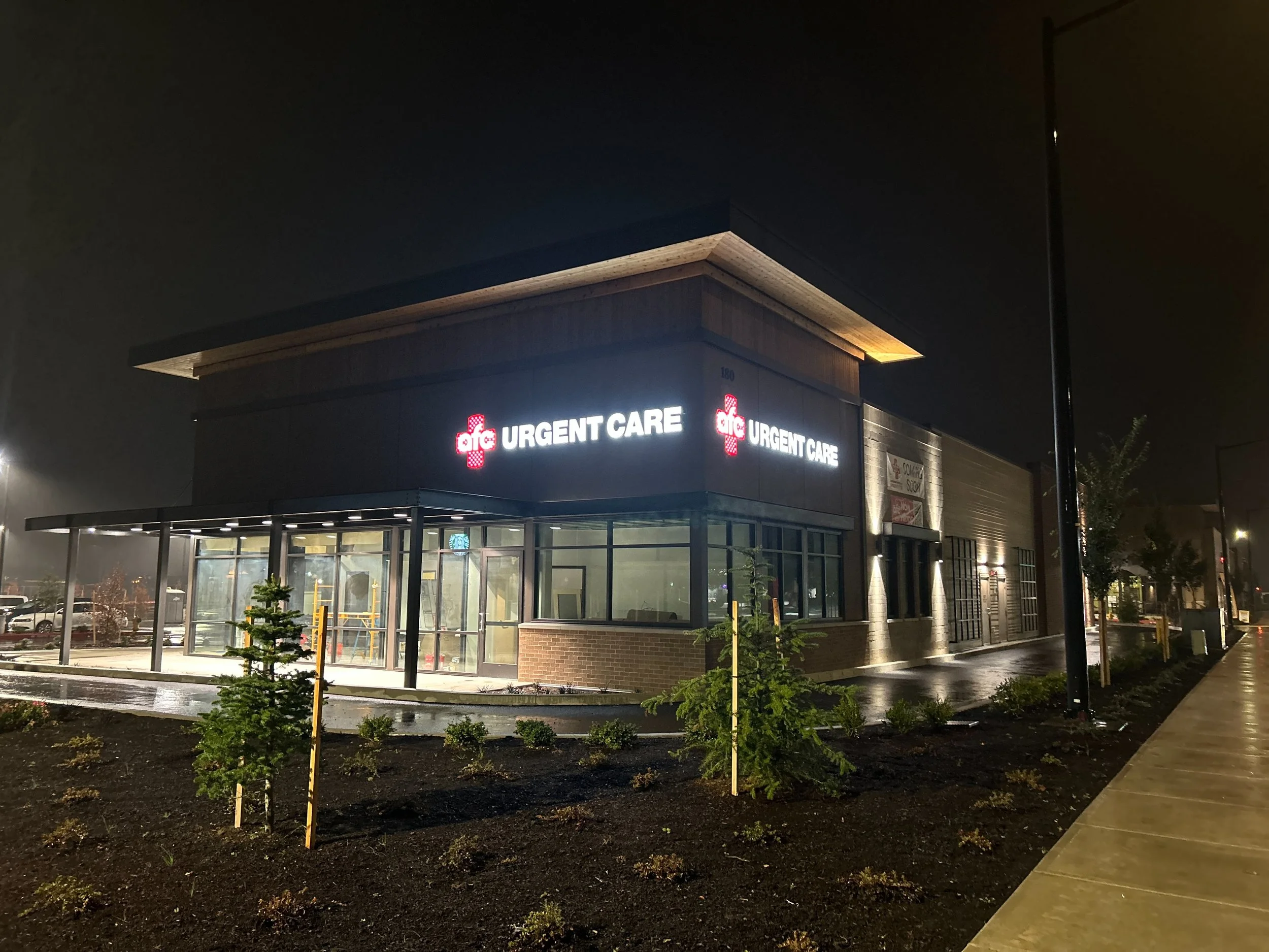 Night view of a modern urgent care building with illuminated sign, glass entrance, and small landscaped area with young trees and bushes.