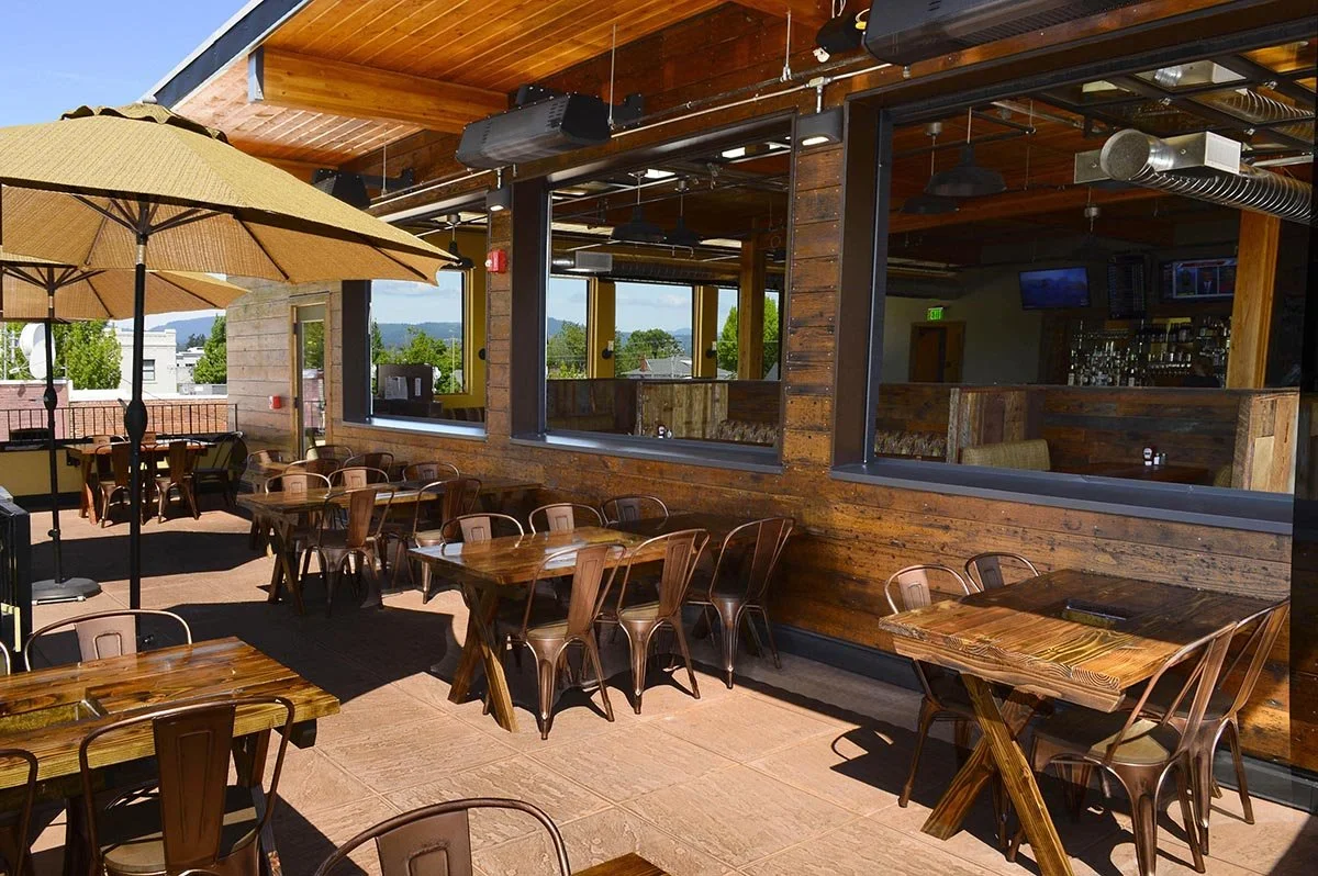 Outdoor patio with wooden tables and chairs, large tan umbrella, and a view of trees and mountains in a sunny day.
