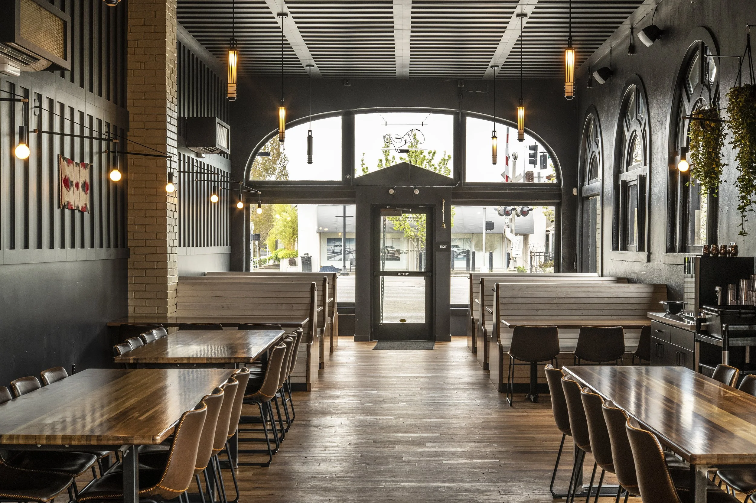 Interior of a modern restaurant or café with wooden tables and chairs, black walls with arched windows, hanging pendant lights, and potted plants on the wall near the entrance.