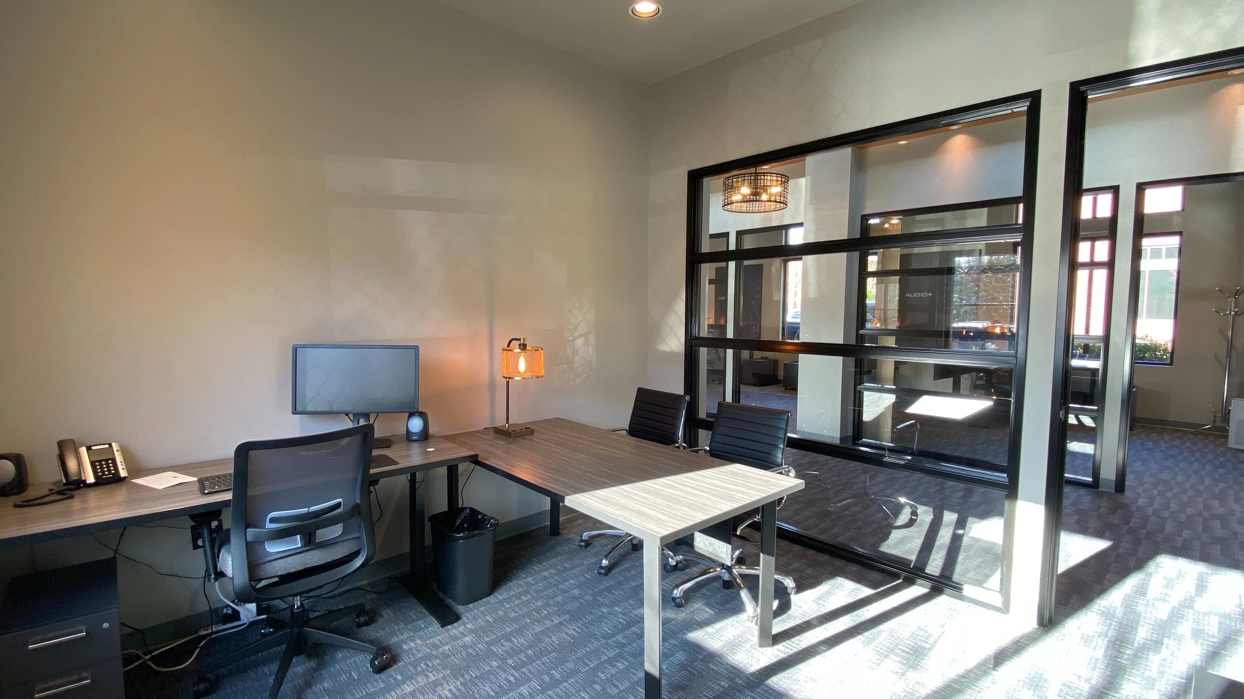 An office with a desk setup. There are two black office chairs, a computer monitor, and two telephones on the desk. A small table lamp is turned on, illuminating part of the workspace. Sunlight is coming through glass doors and windows, casting shado