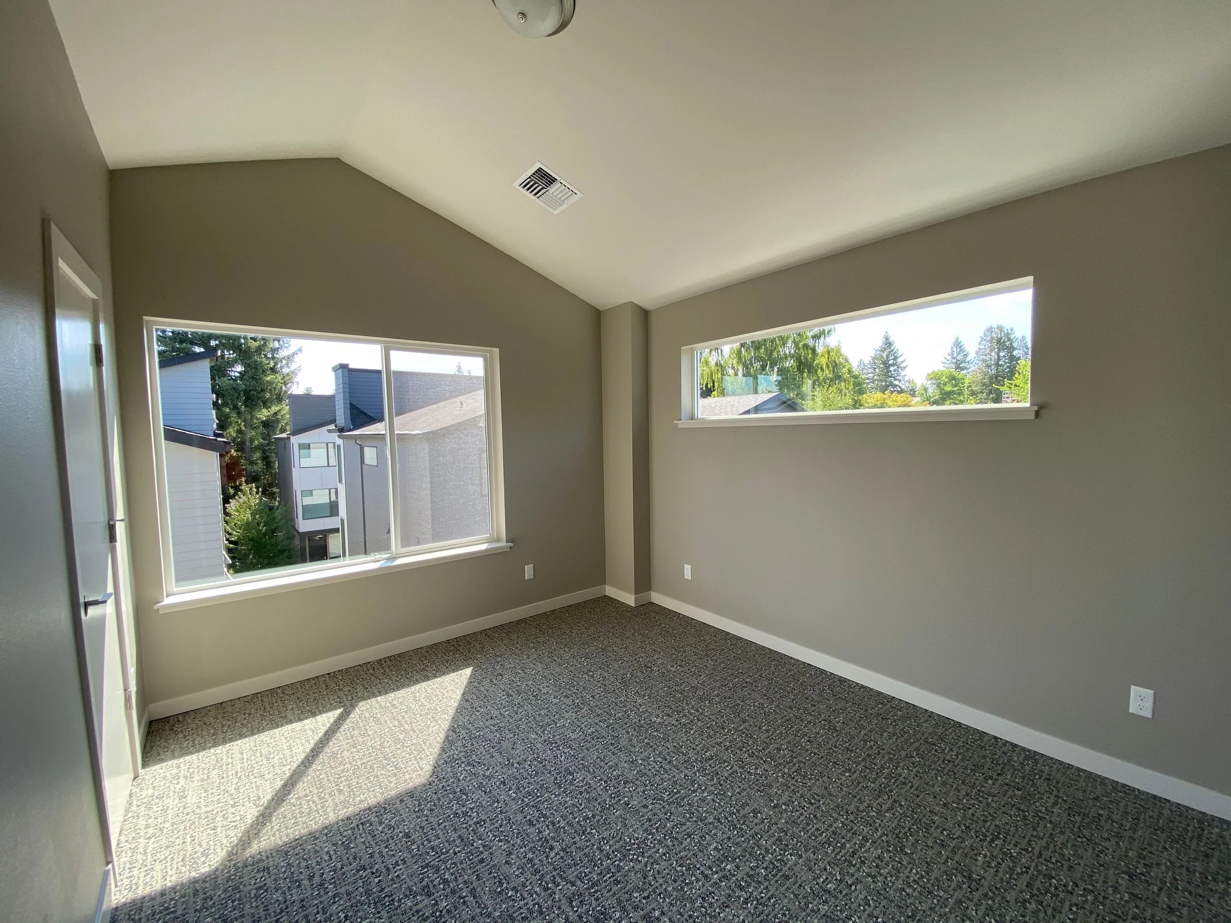 Empty room with two large windows, neutral-colored walls, gray carpet, and a ceiling light fixture.