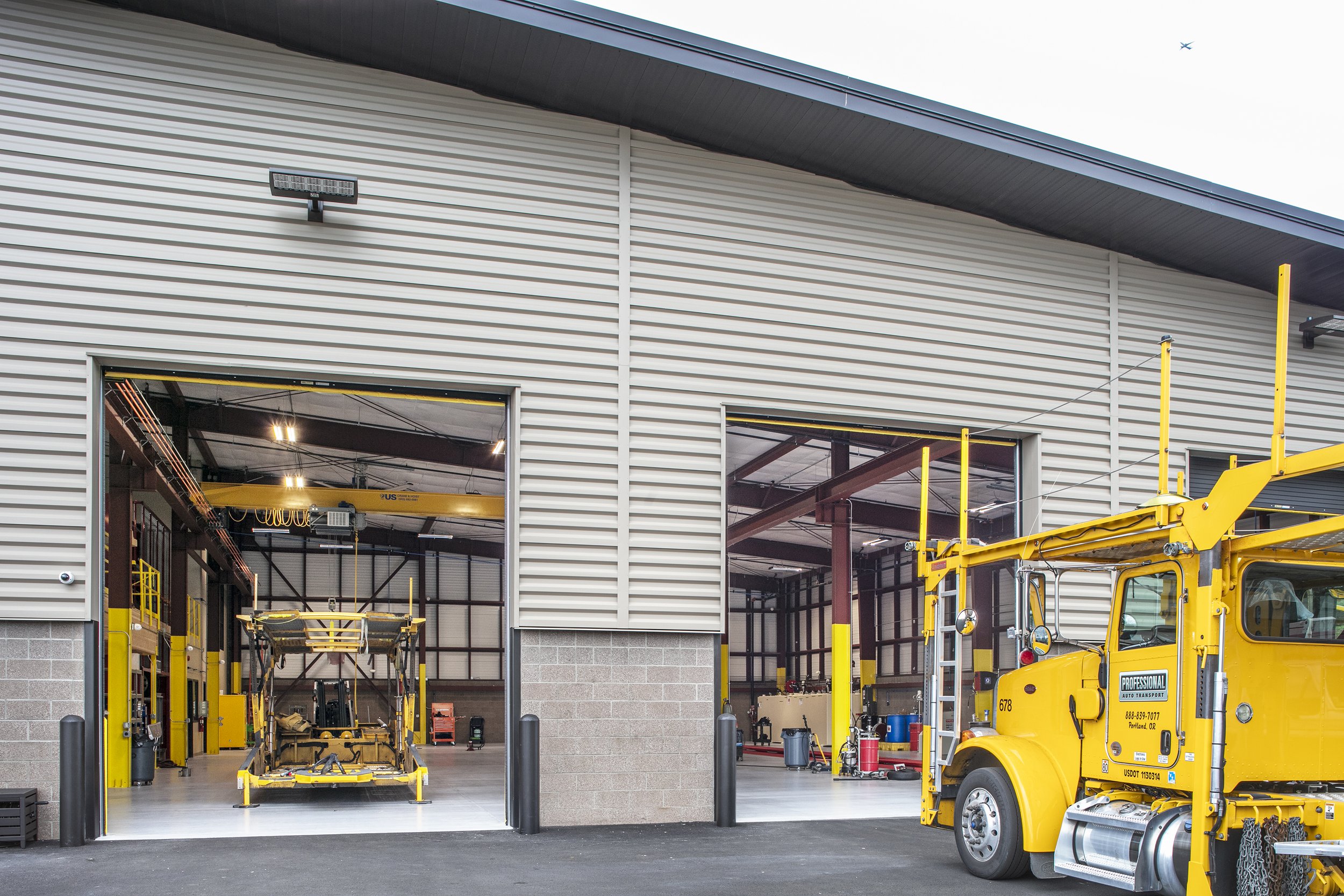 Inside a vehicle repair shop with a yellow tow truck parked outside and a yellow vehicle inside the garage bay.