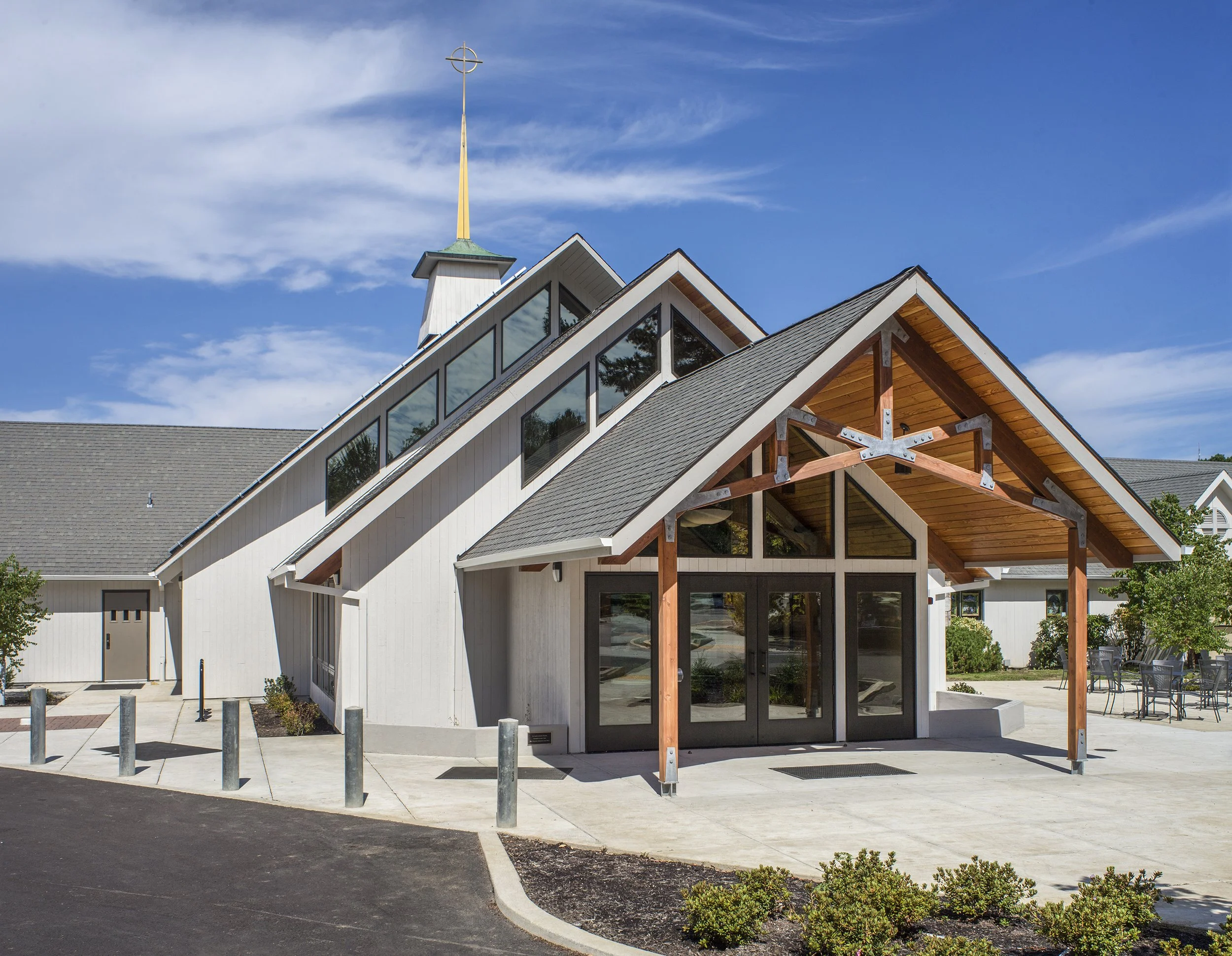 Modern church building with large glass windows, wooden accents, and a steeple, surrounded by parking area and outdoor seating, under a blue sky.