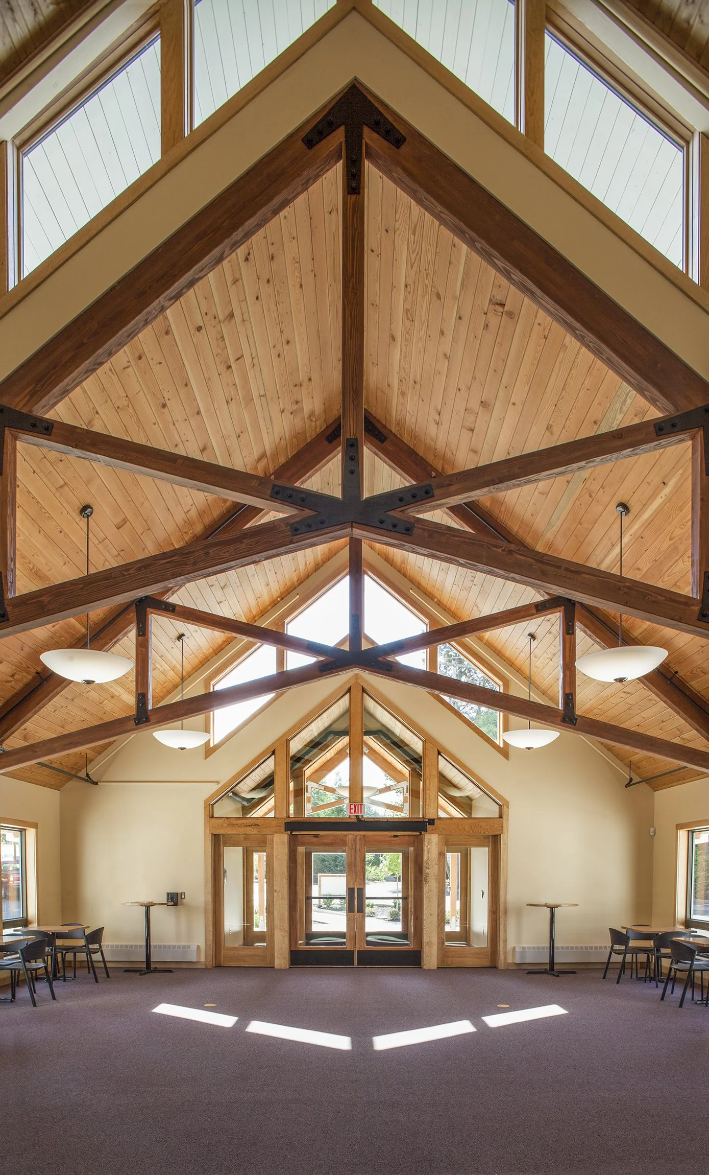 Interior of a spacious room with a high, vaulted wooden ceiling and large triangular windows allowing natural light, with tables and chairs along the walls.