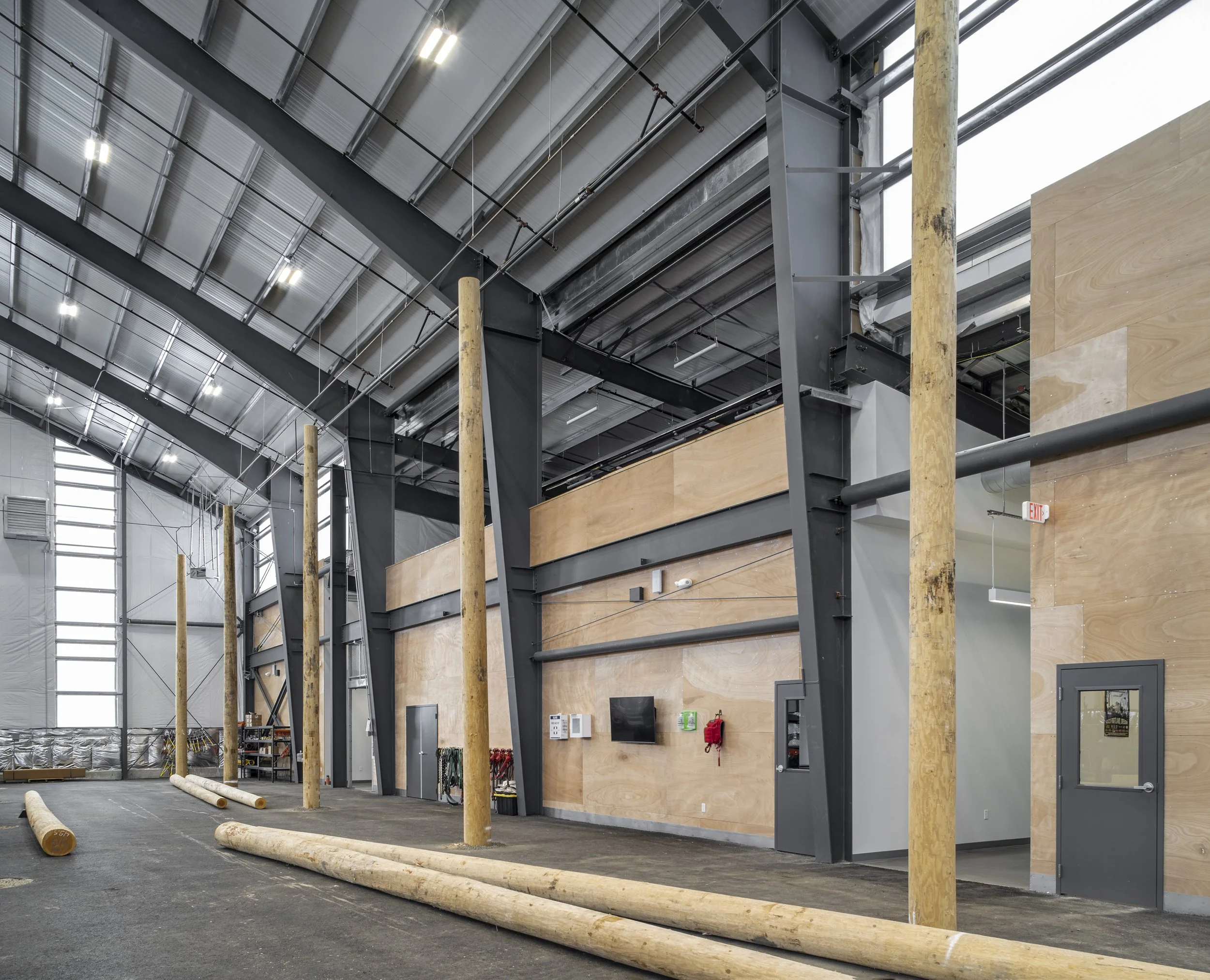 Interior of a warehouse or gym with wooden poles on the floor, a high ceiling with lights, and a partial wooden wall with safety equipment and a monitor.