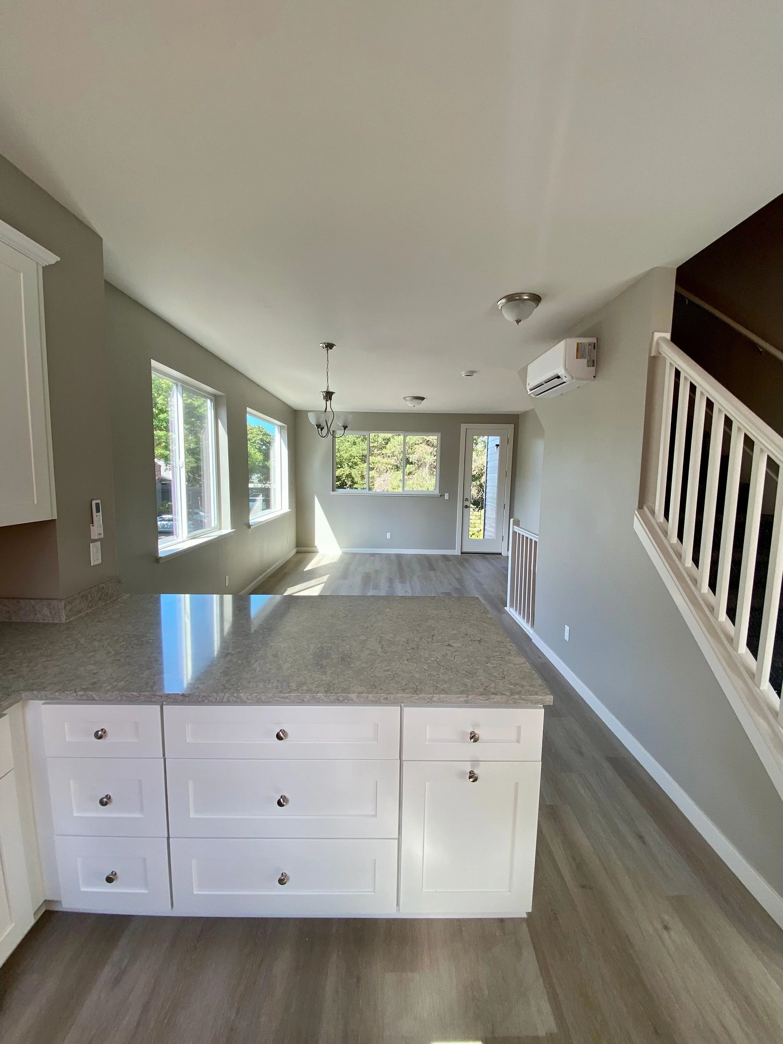 View of an open kitchen and living room area with large windows, white cabinetry, granite countertop, and hardwood floors, with sunlight streaming in.
