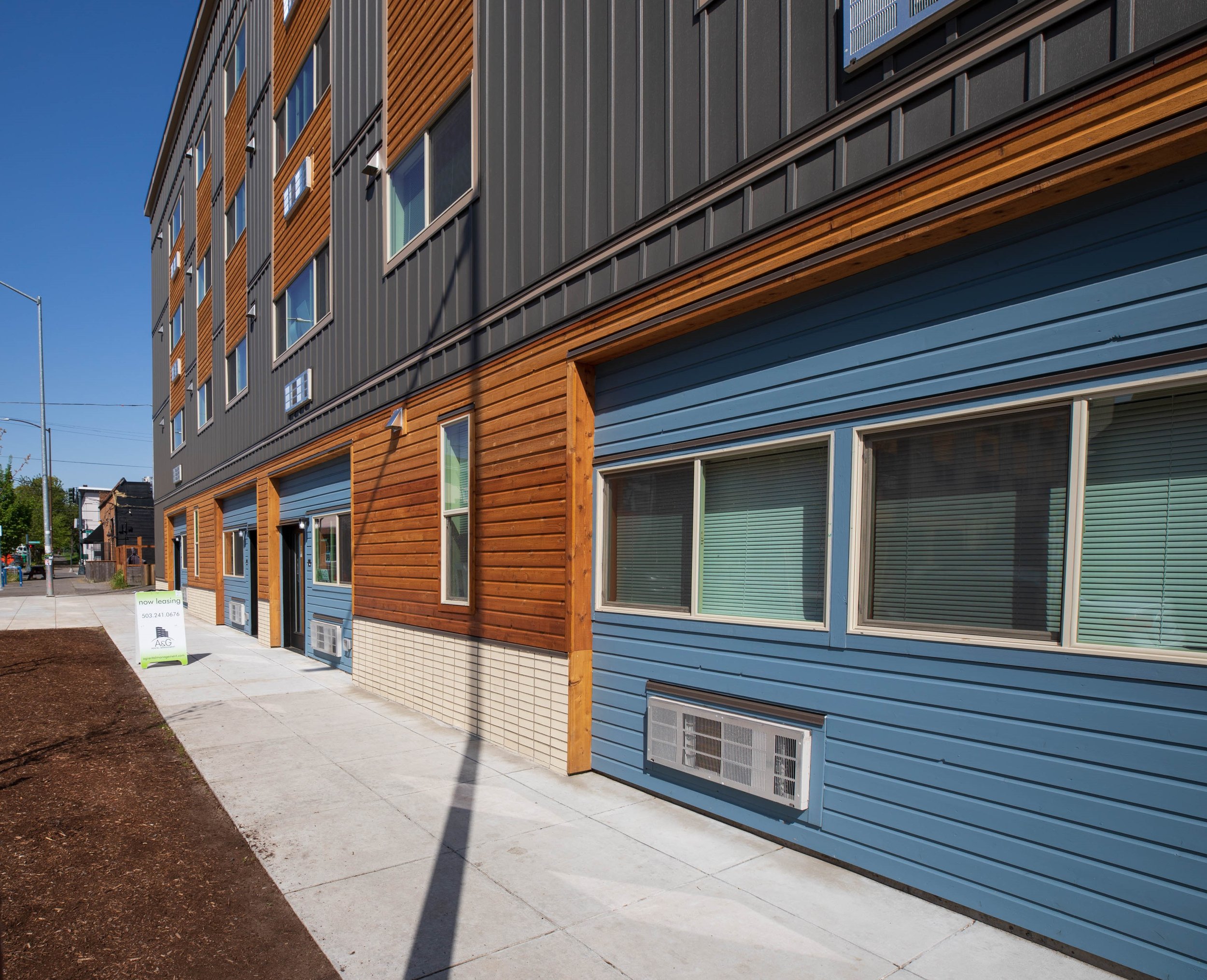 A modern apartment building with wood, black, and blue siding, multiple windows, and several air conditioning units on the lower wall. There is a sidewalk and a small patch of dirt in front of the building under a clear blue sky.
