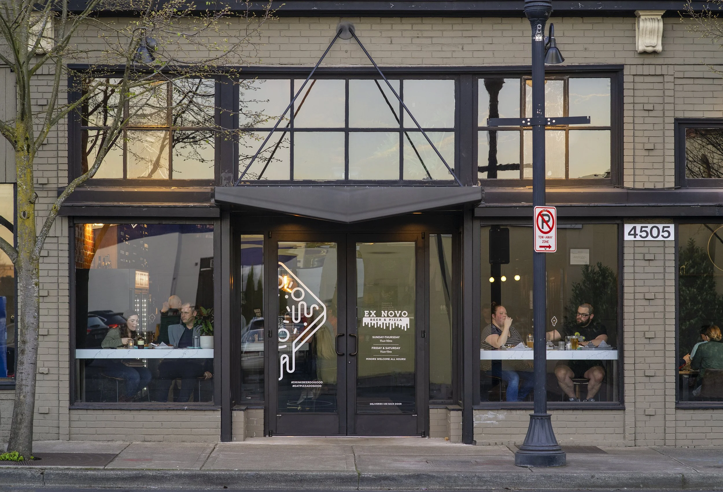 Front view of a restaurant with large glass windows, a glass door, and brick exterior, showing people dining inside. The restaurant has a building number 4505, and a street lamp and no parking sign outside.