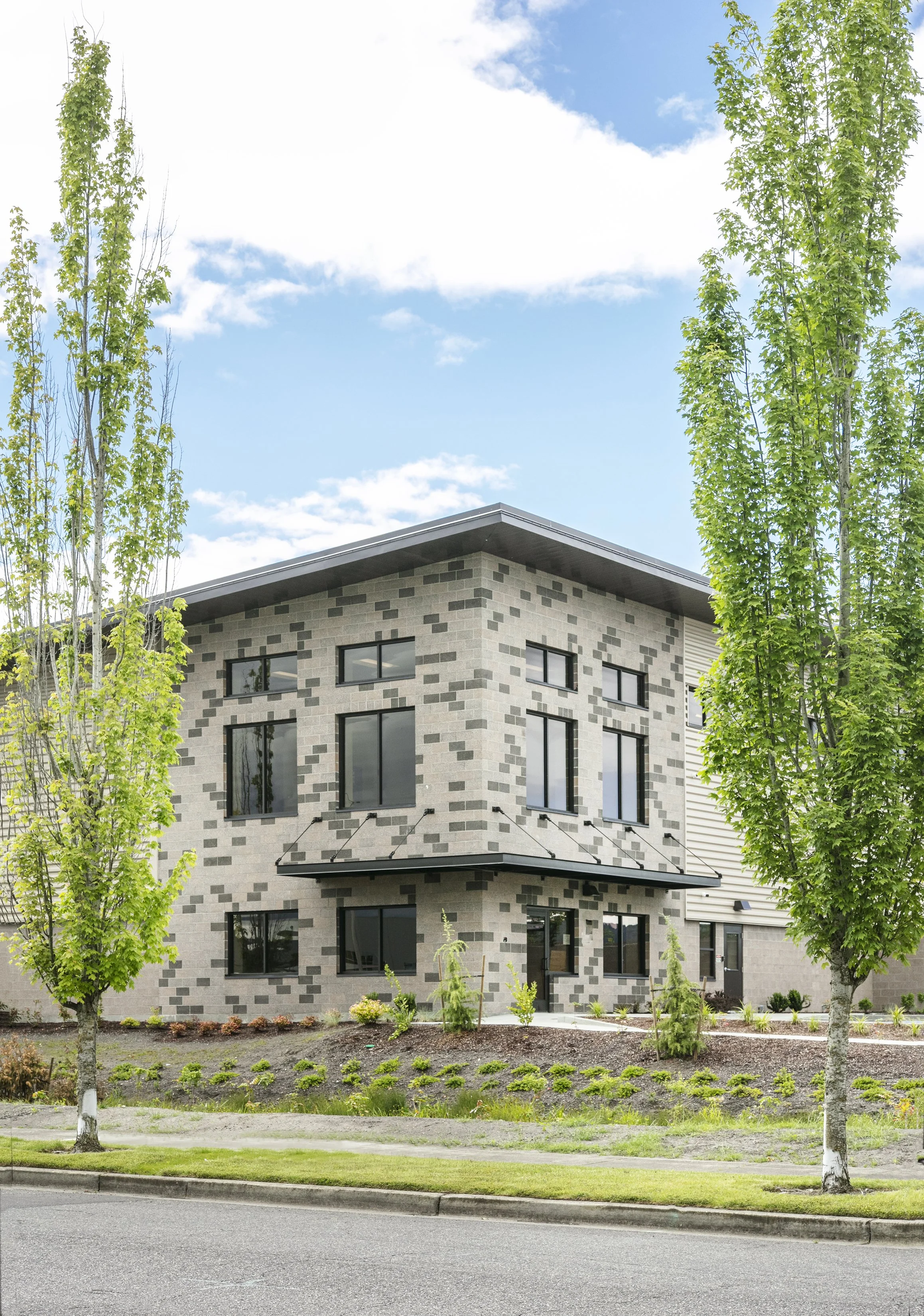 A modern three-story residential building with a gray brick exterior, black-framed windows, and a small front yard with green plants and trees, on a partly cloudy day.