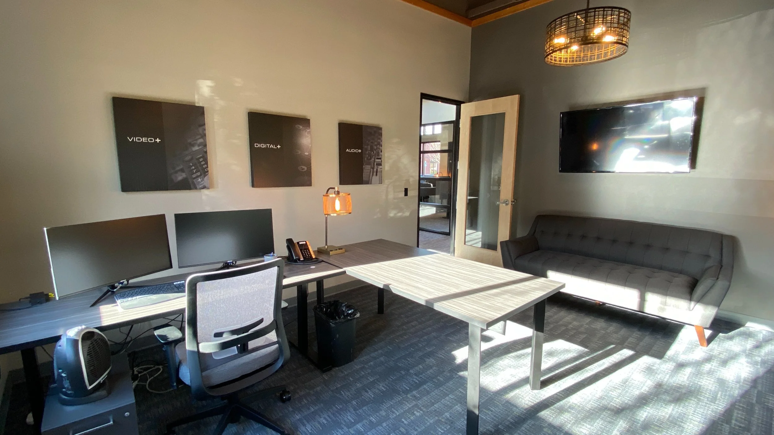 Office with a desk containing two computer monitors, a lamp, a telephone, and a trash bin. There is a gray couch, a TV on the wall, and three framed posters about media and digital topics. A door is open to another room with large windows.