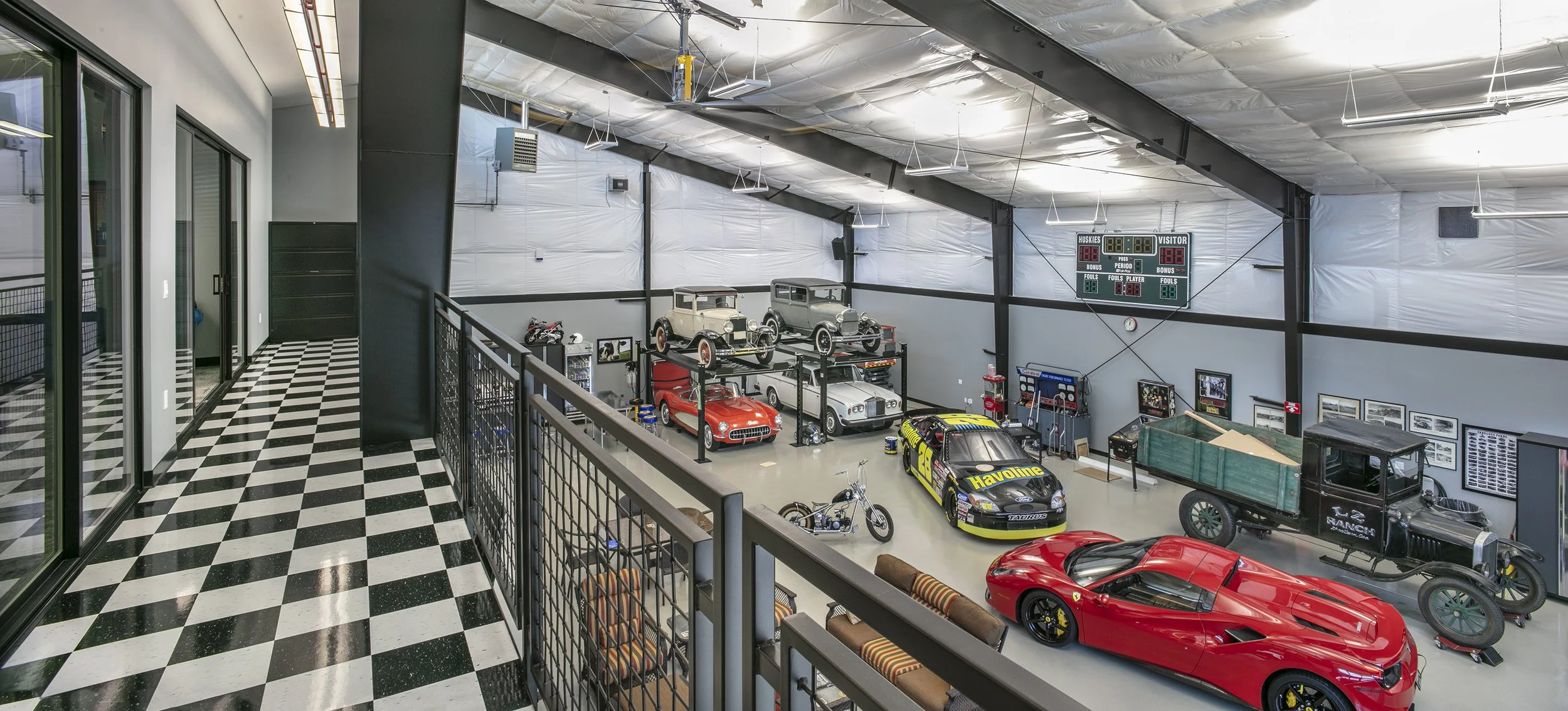 Indoor garage or museum exhibition with vintage and modern cars, motorcycles, and car memorabilia. There are classic cars on display, a racing car with sponsorship decals, and various automotive artifacts along the walls. The space has high ceilings 