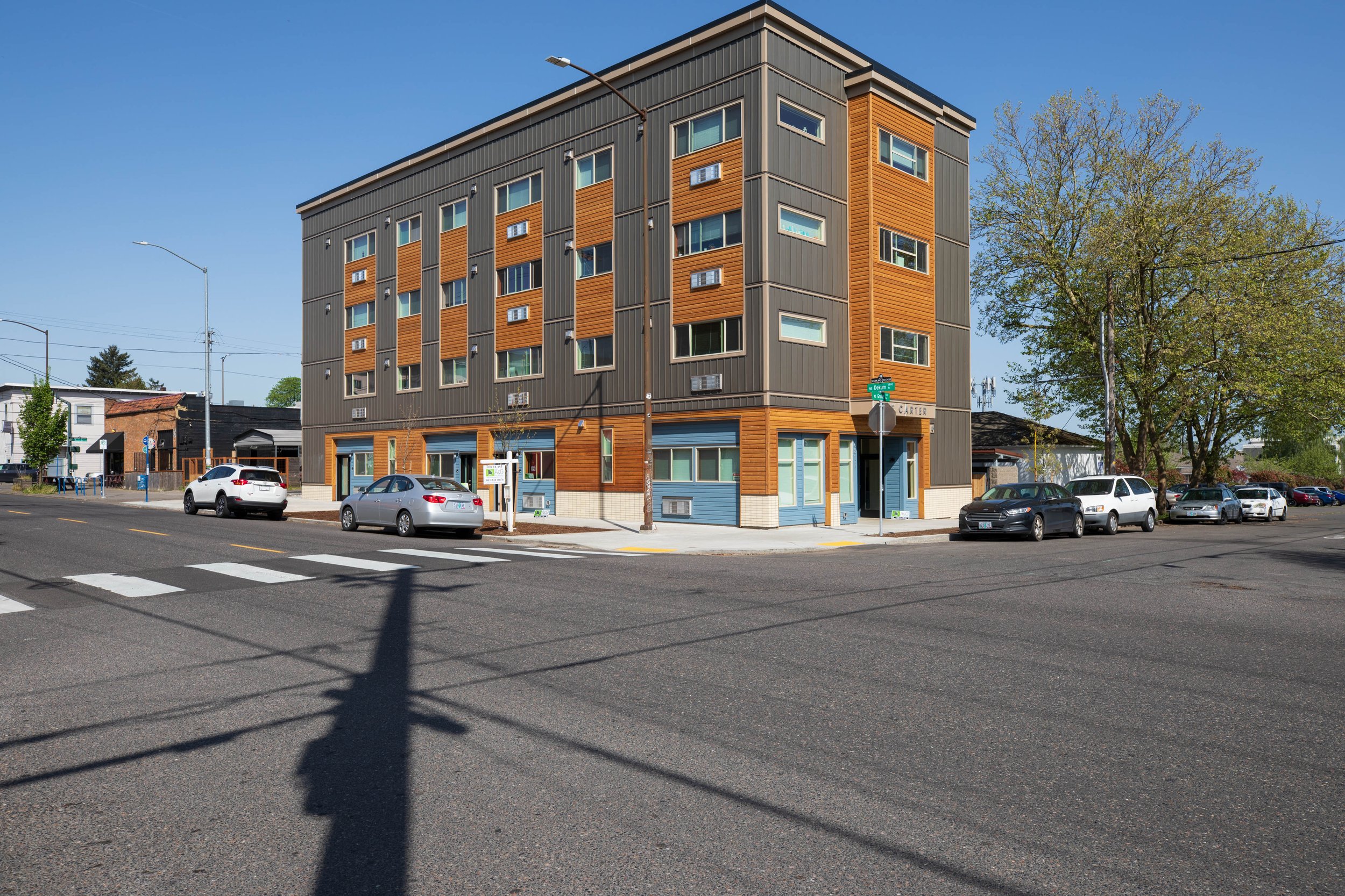 A four-story mixed-use building with commercial space on the ground floor and residential units above, located on a street corner with parked cars and a crosswalk in front, under a clear blue sky.