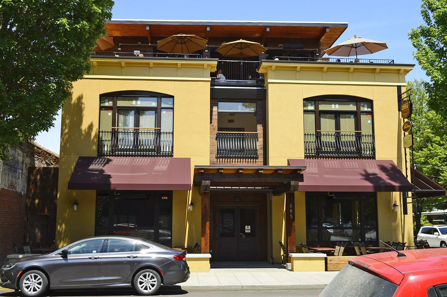 A three-story building with a yellow facade, black railings, and large windows. The ground floor has a restaurant or cafe with outdoor seating, maroon awnings, and a central entrance with wooden support beams. The upper floors have balconies with umb