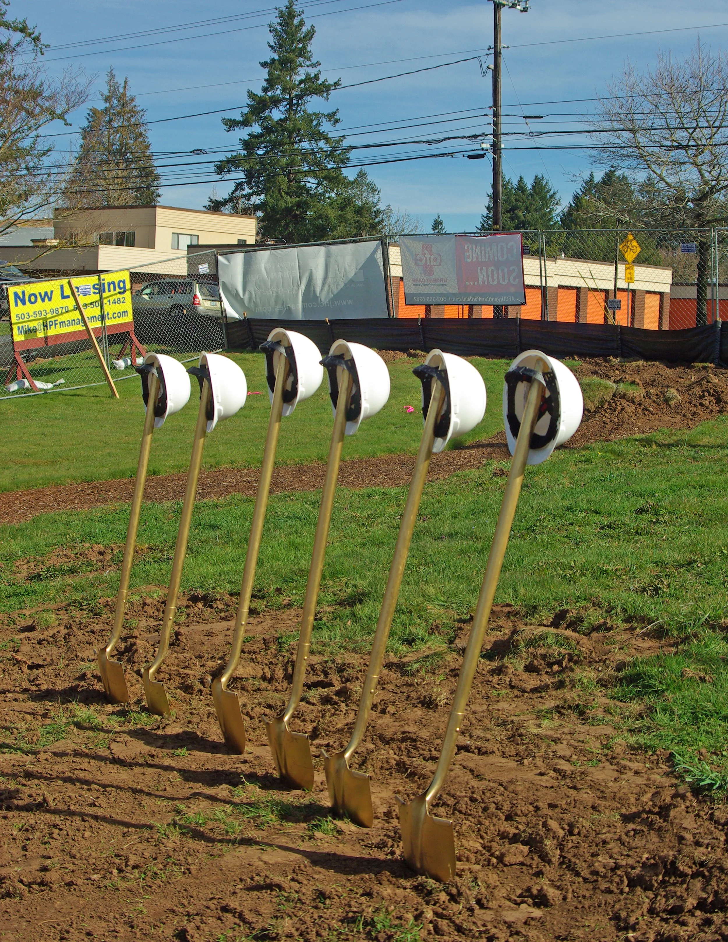 Six shovels with white helmets hanging from their handles, planted in the ground in a row at a construction or groundbreaking site, with grass and dirt in the foreground, and a construction fence, trees, and buildings in the background.