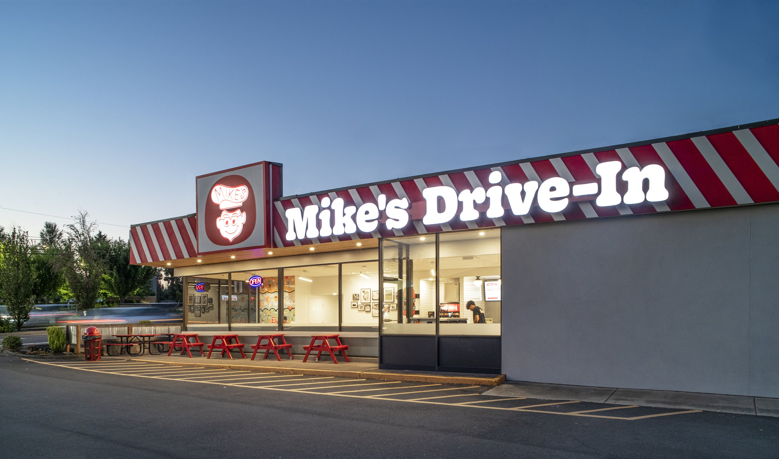 Exterior view of Mike's Drive-In restaurant with a large illuminated sign, red and white striped awning, and red picnic tables outside during dusk with trees and a parking lot in the background.