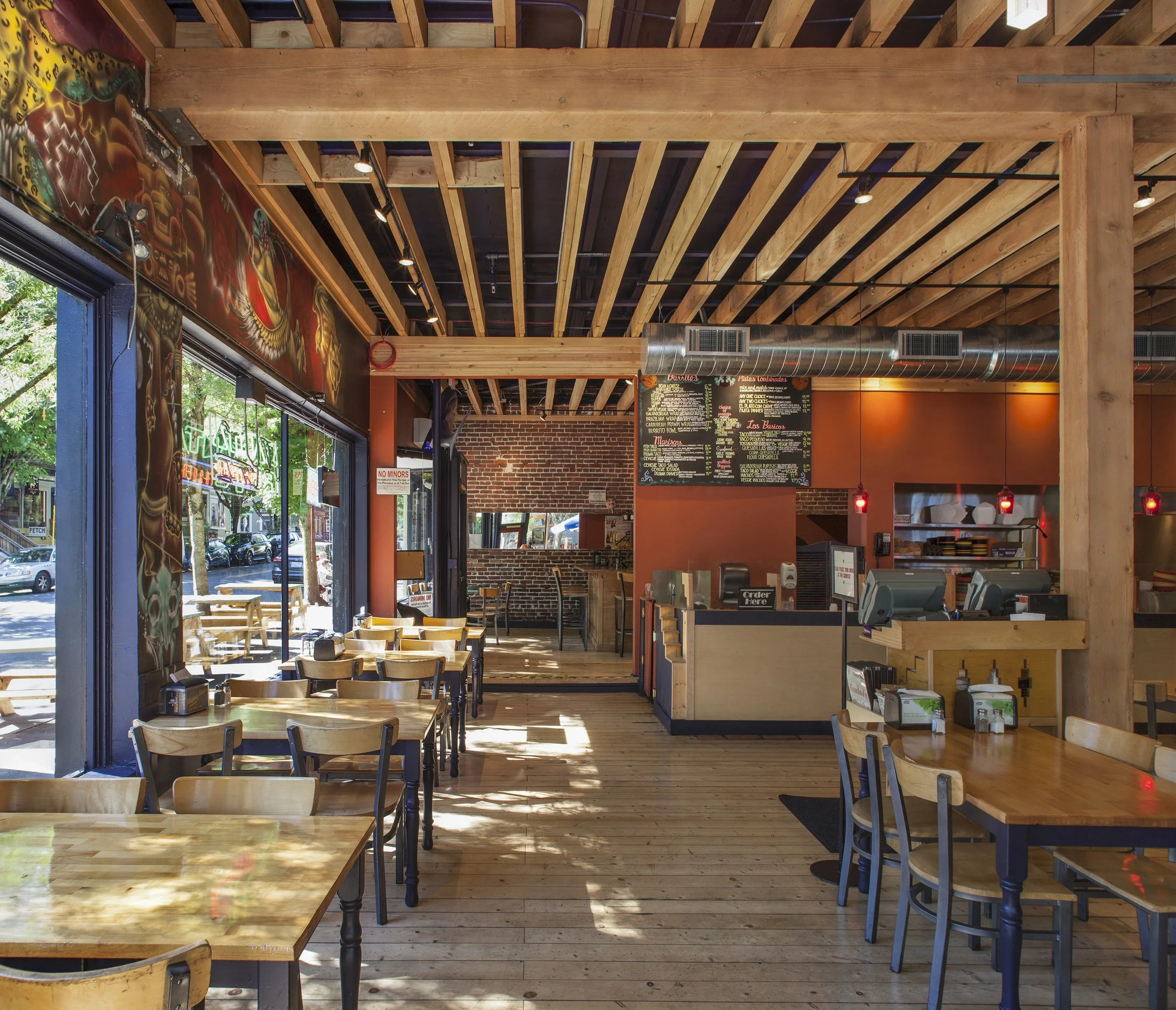 The image shows the interior of a cozy restaurant with wooden tables and chairs. Large windows let in natural light, illuminating the wooden floor. The decor includes colorful wall murals and warm lighting fixtures. A menu board is visible behind the