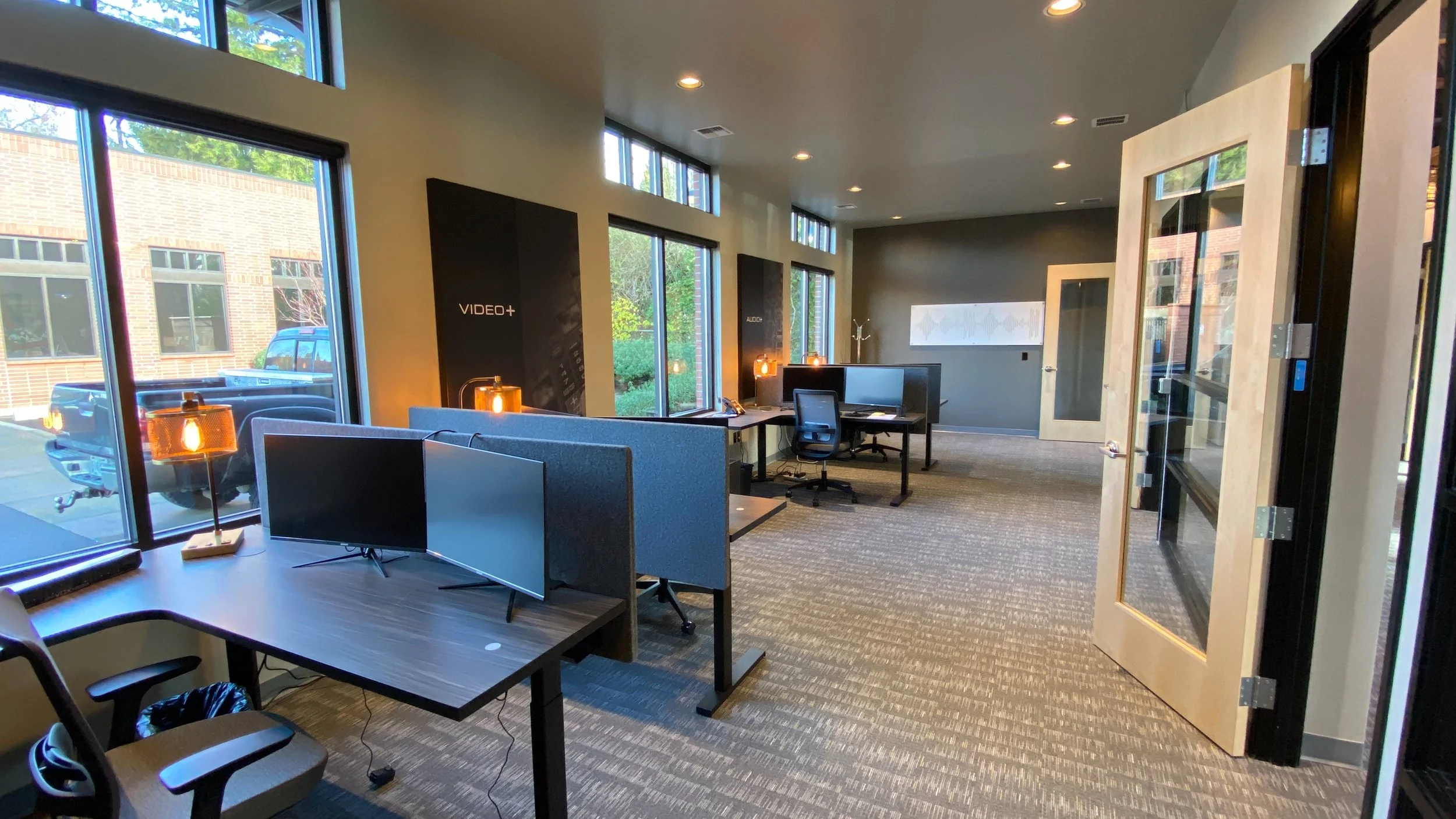 Modern office space with multiple workstations and computers, large windows with outside view, art wall decor, and a glass door entrance.