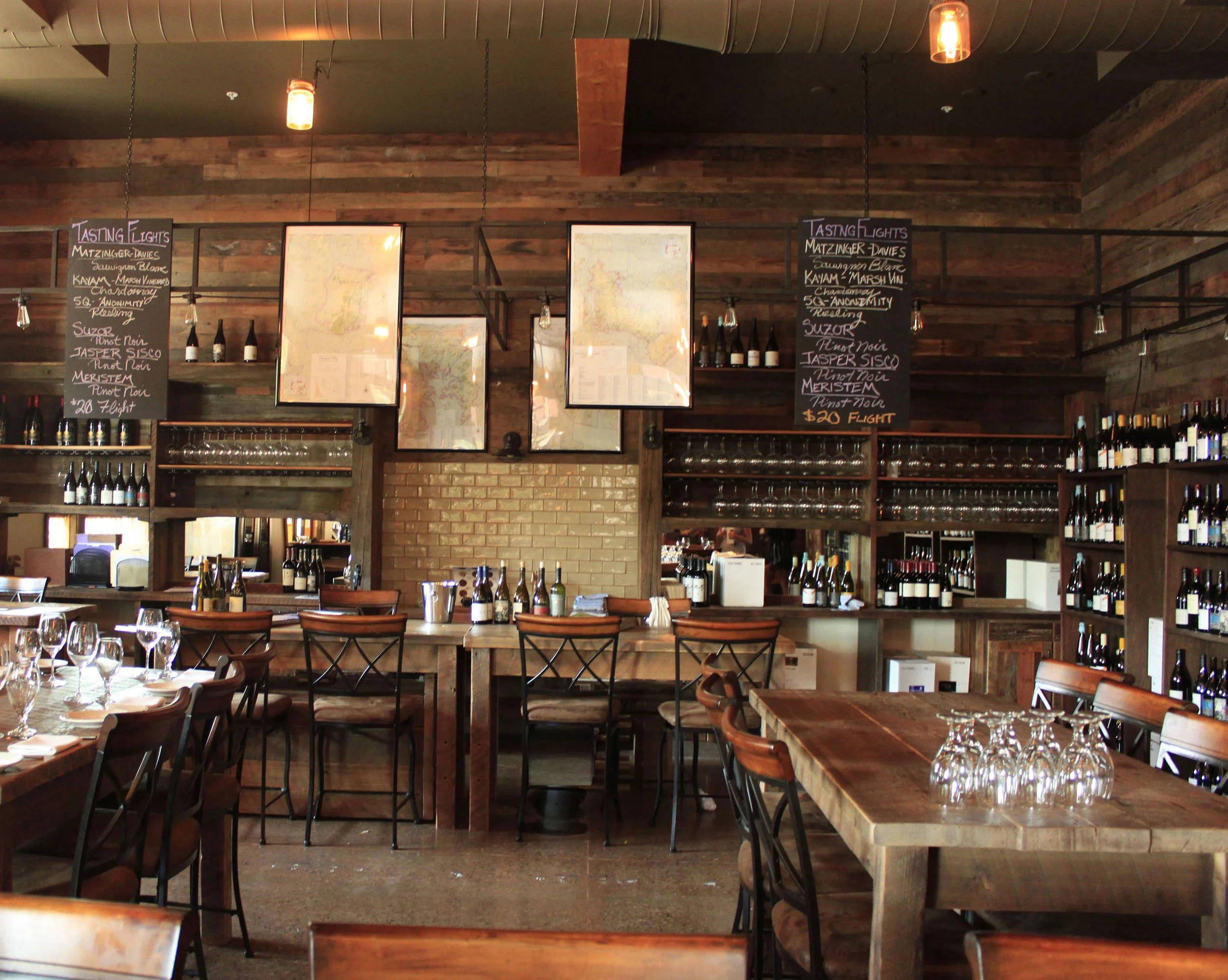 Interior of a restaurant with wooden tables and chairs, wine bottles on shelves, chalkboard menus, and hanging pendant lights.