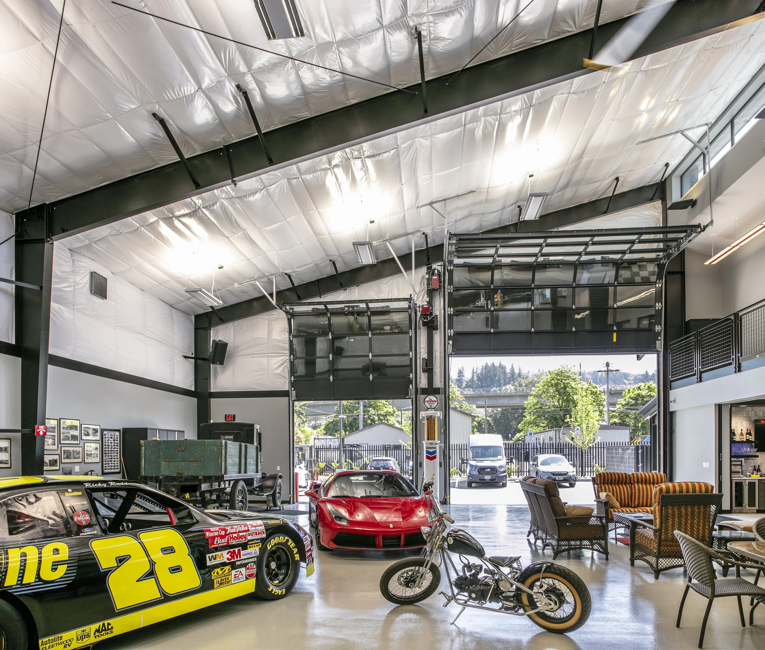 Indoor showroom with a red race car, vintage motorcycle, muscle car, motorcycle, and seating area, with open garage doors revealing parked vehicles outside.