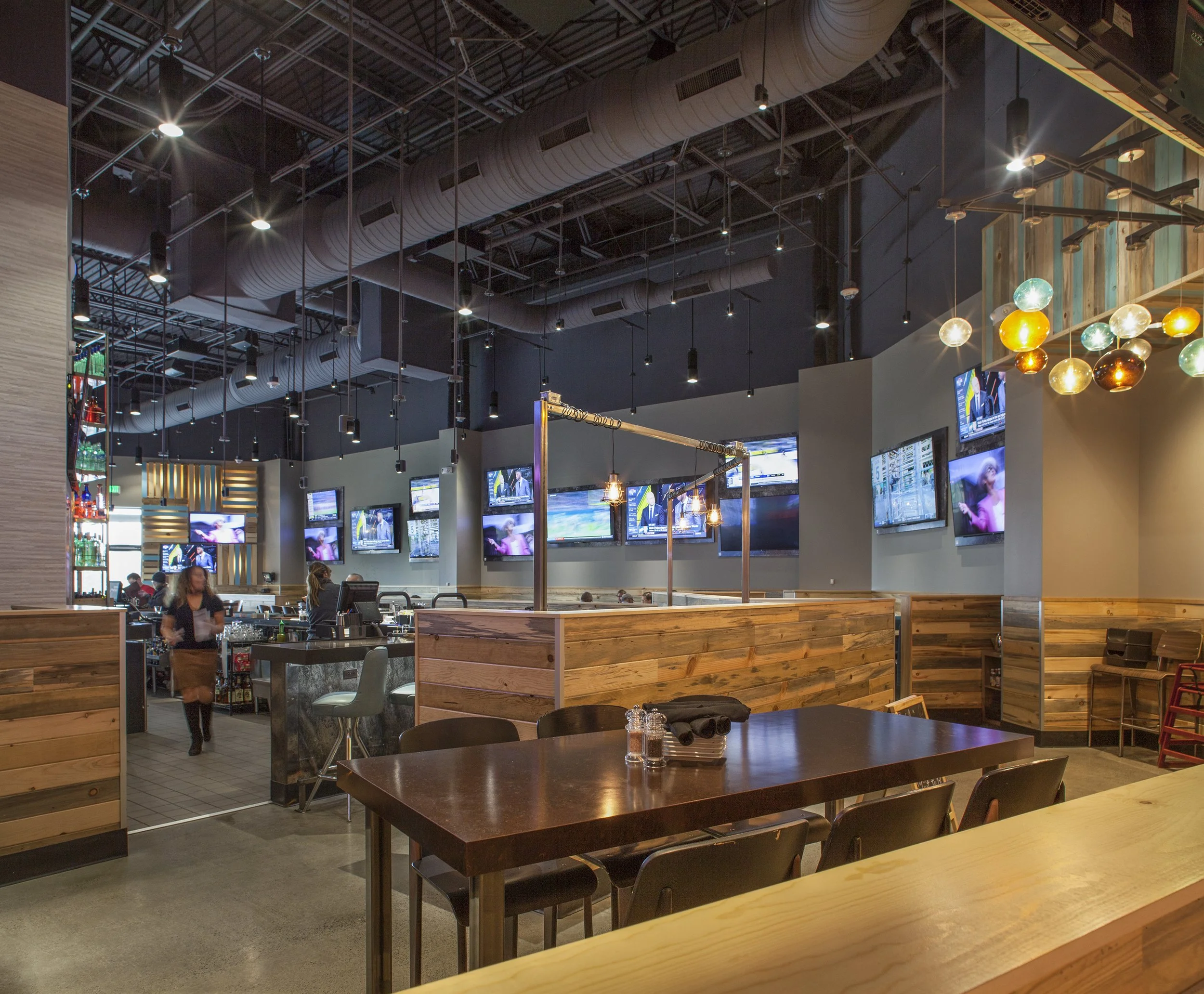 Interior of a modern sports bar with wooden accents, multiple flat-screen TVs on the walls, a bar counter with barstools, and decorative hanging lights.