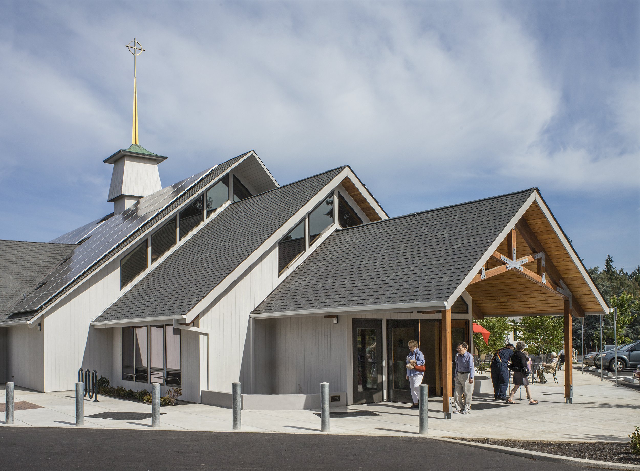 Modern church building with solar panels on roof, wooden entrance with people entering and outdoor seating area.