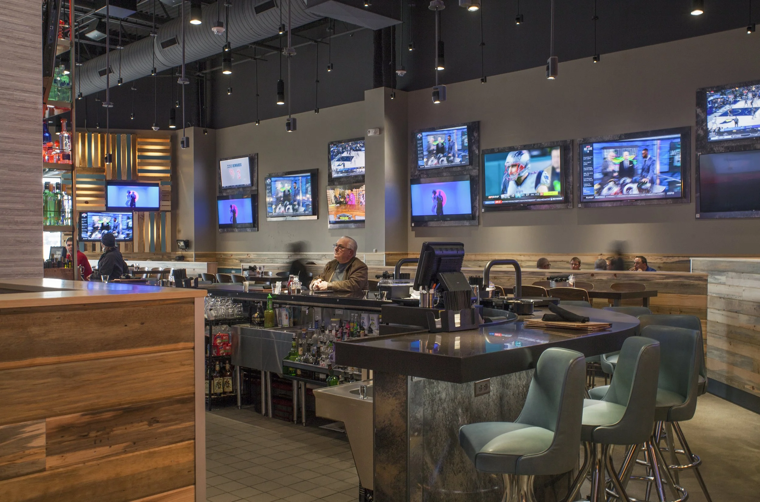 A modern bar and sports lounge with multiple television screens displaying sports events. The bar area has chairs and a bartender, with a wooden counter and shelves of bottles. There are patrons sitting and watching TV, some seated at tables in a coz