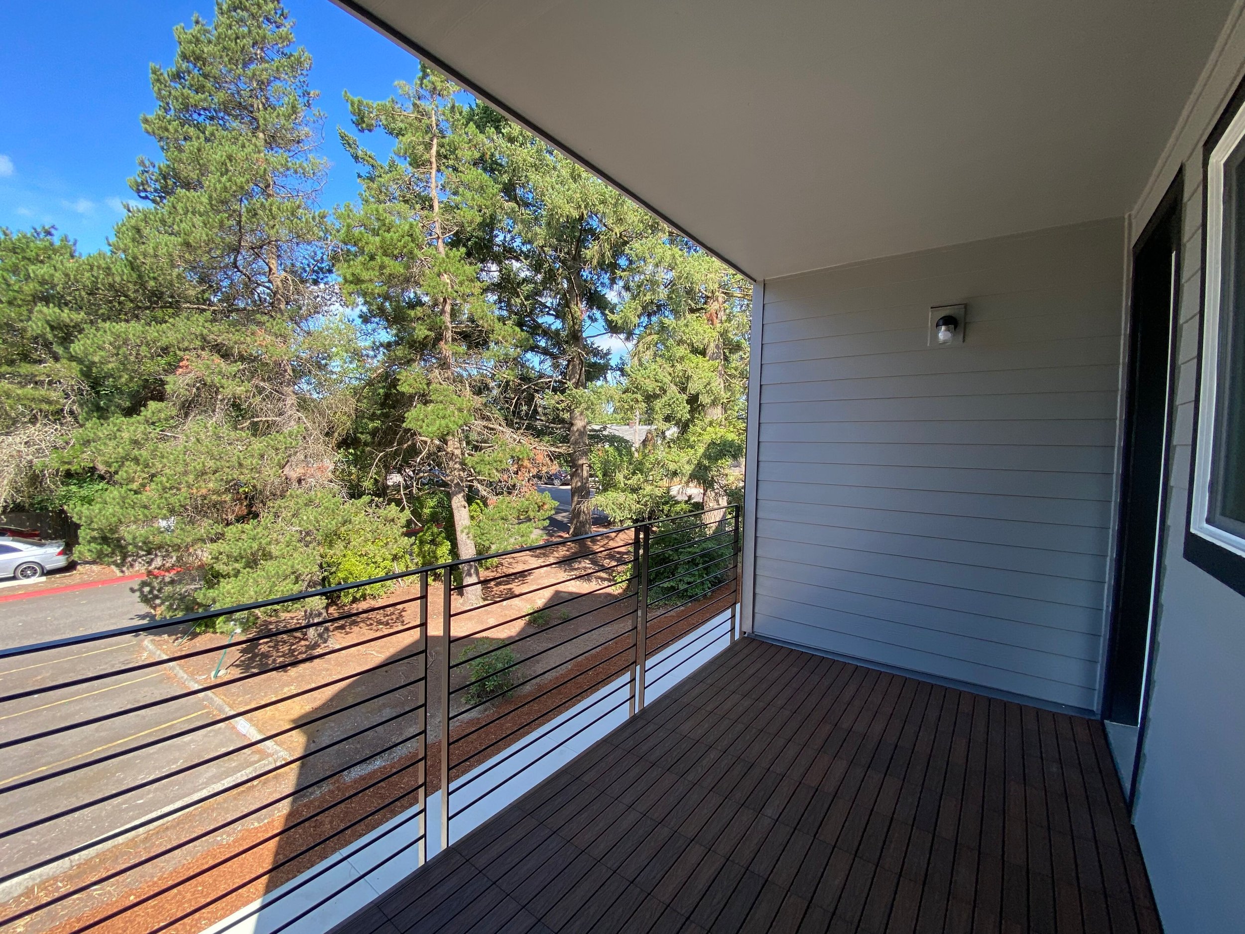 Balcony with wooden floor, gray siding wall, metal railing, and view of trees and parked cars outside.