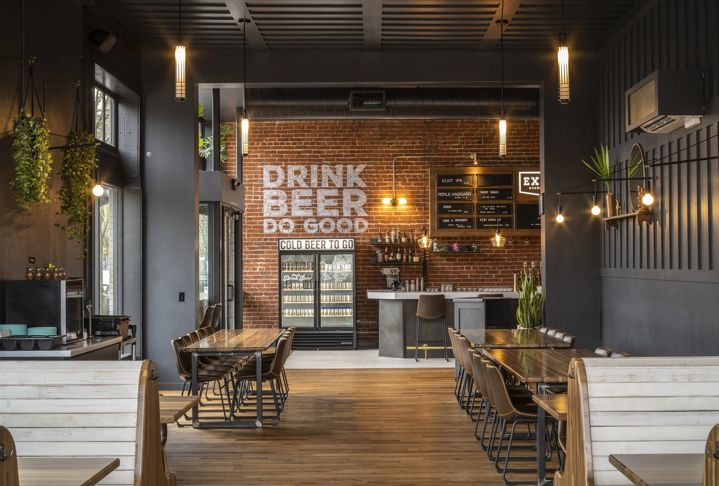Interior of a modern brewery or bar with brick walls, wooden floors, and seating area. A large sign on the brick wall says 'DRINK BEER DO GOOD.' There is a refrigerator with cold beer to go, a bar counter with bottles and glasses, and a menu board wi