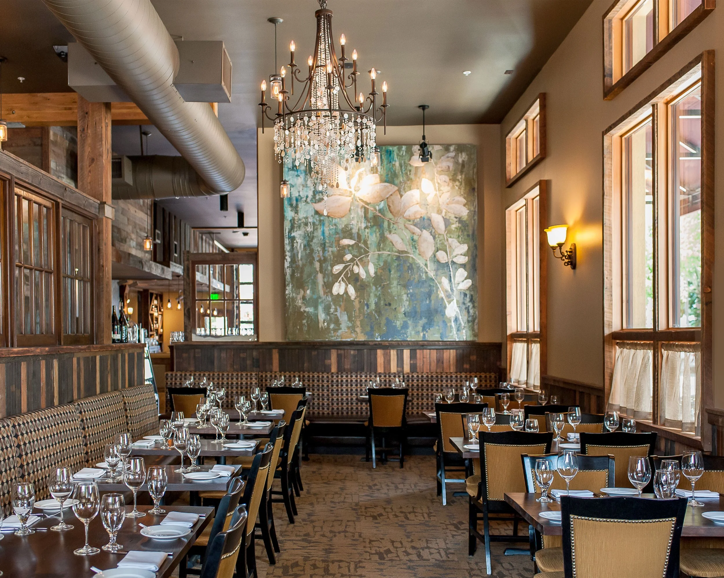 Empty restaurant dining area with tables set for service, featuring wine glasses, napkins, and cutlery, large windows, a chandelier, and a wall with a large floral painting.