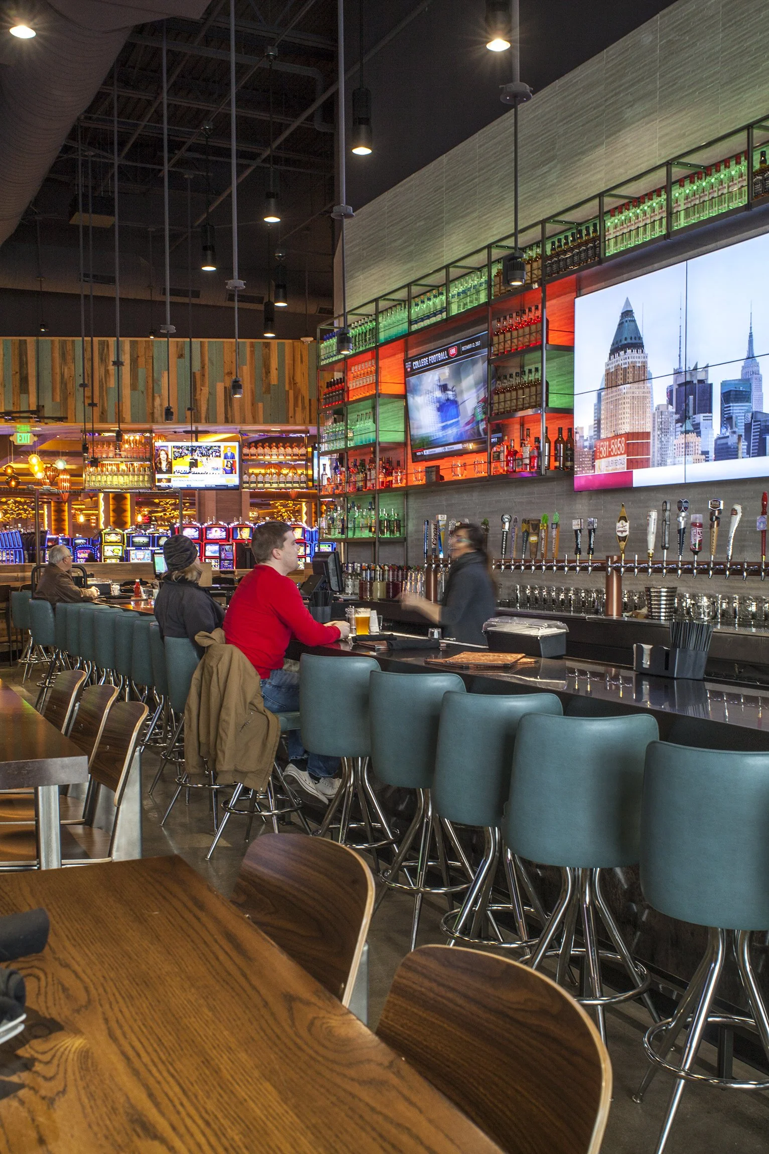 A lively sports bar with wooden and teal bar stools, a long bar counter with various beer taps, large TV screens showing cityscapes and sporting events, colorful illuminated shelving of alcohol bottles, and a few patrons seated at the bar. In the bac